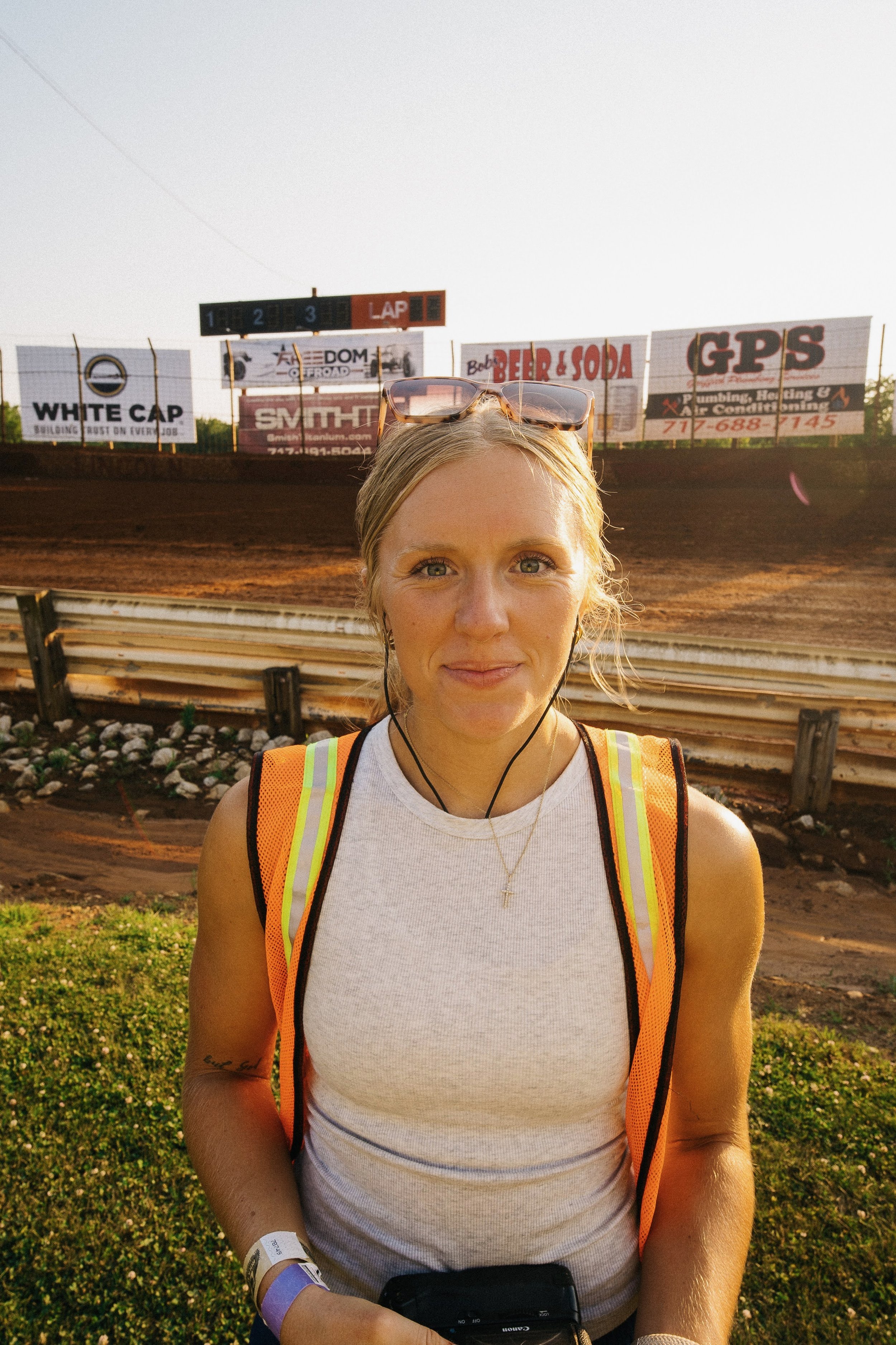 A young woman with blonde hair and sunglasses on her head, wearing a white tank top and yellow safety vest, standing in front of a dirt race track with advertising banners.