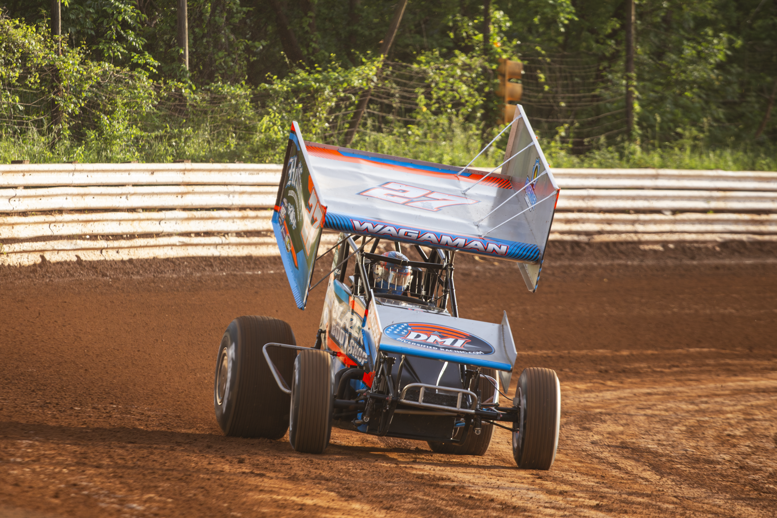 A sprint car racing on a dirt track with a green forest background and a wooden barrier.