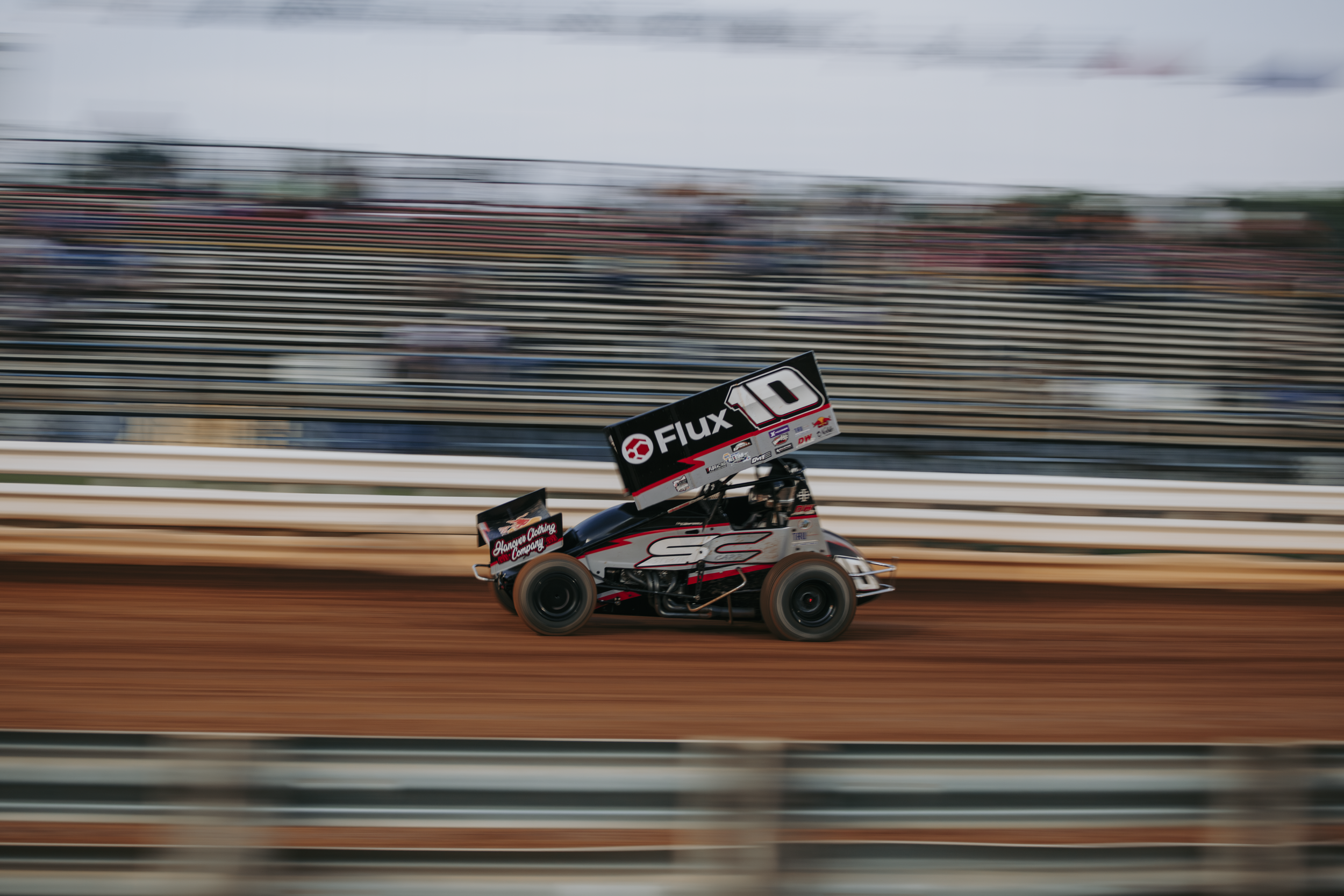 A sprint car racing on a dirt track with a blurred background and a large black wing with white and red lettering.