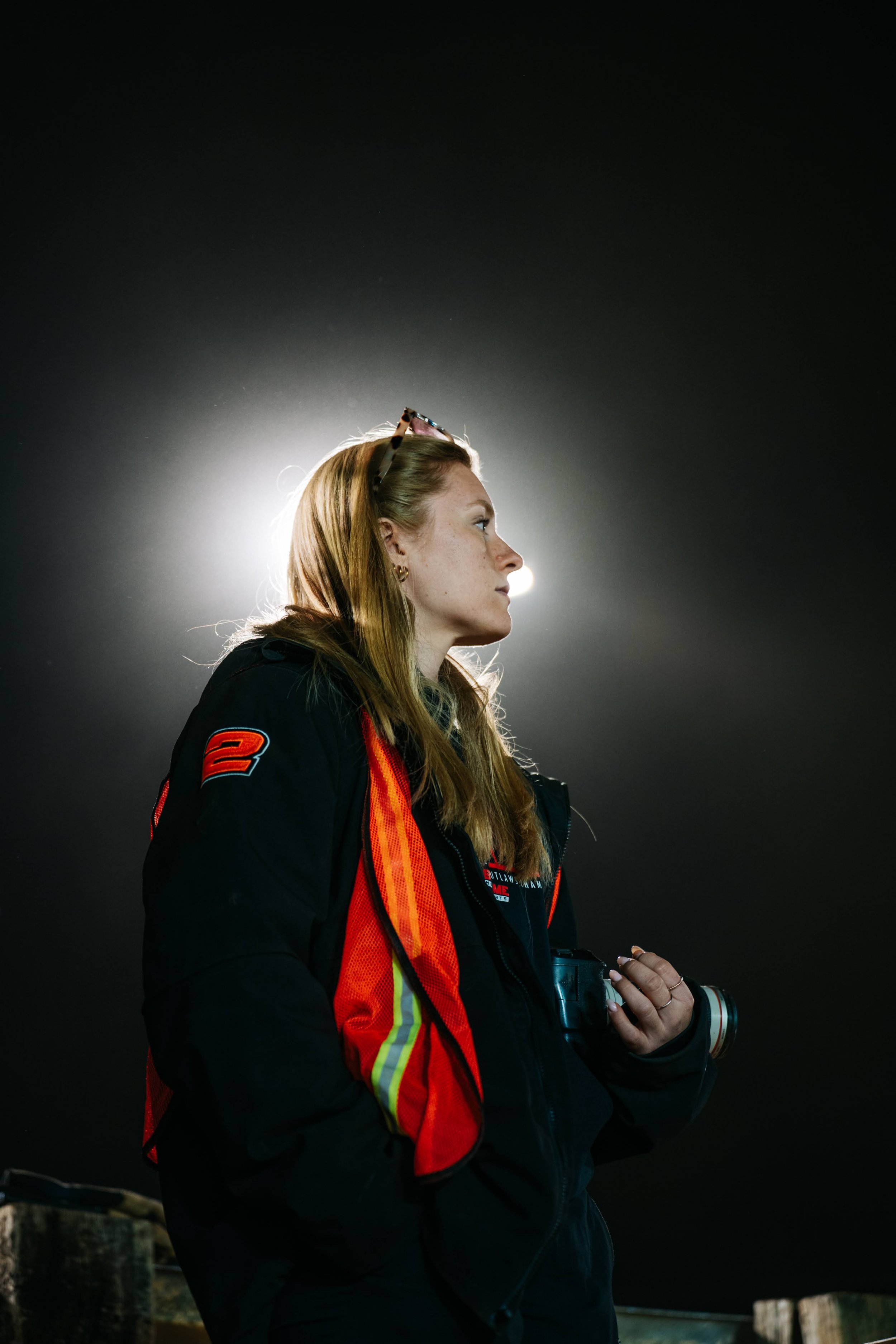 Profile of a woman with red hair, wearing a black jacket with orange and yellow reflective stripes, holding a camera, and sunglasses on her head, against a dark background with bright light behind her.