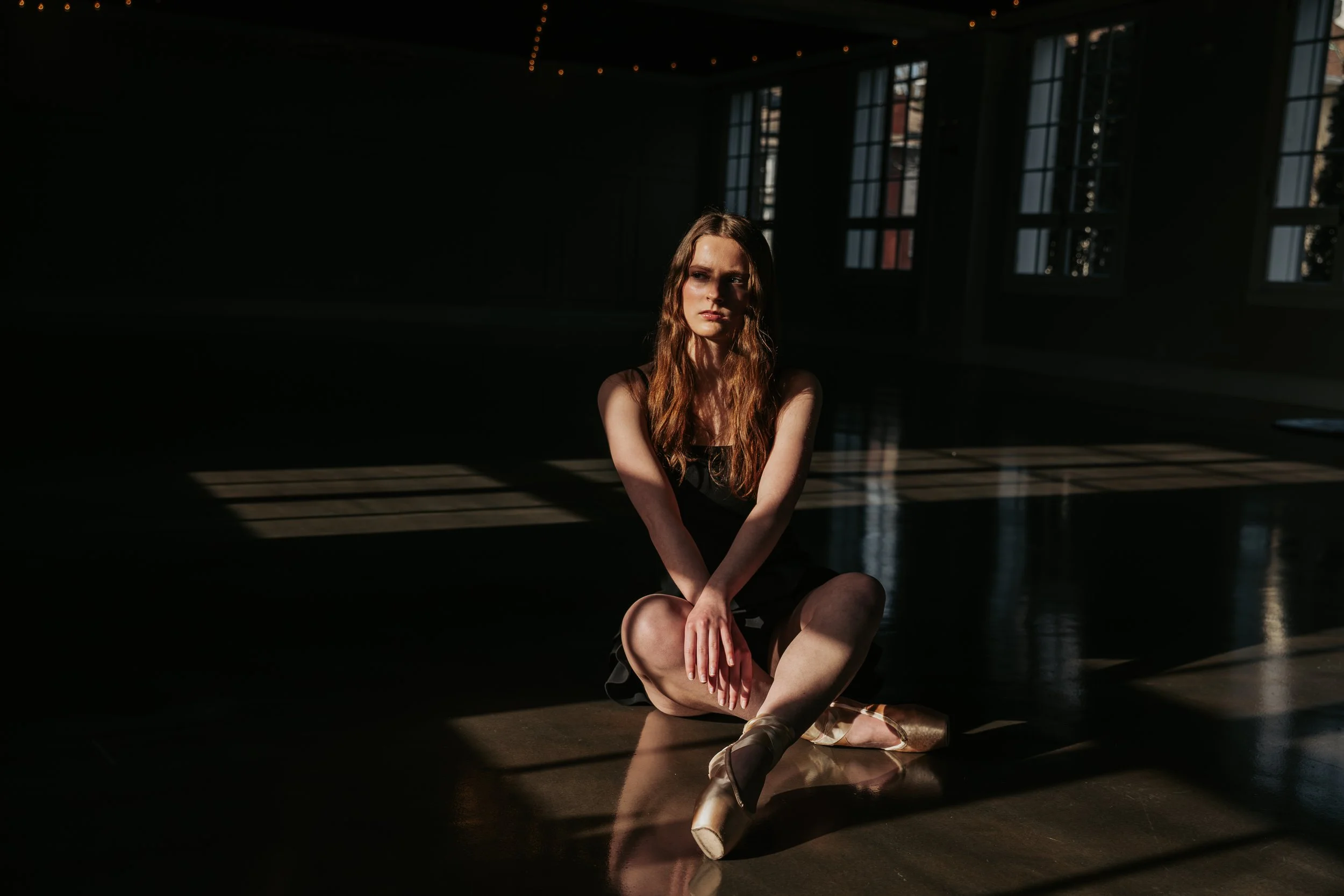 A young woman with long wavy hair sitting on a ballet studio floor with sunlight streaming through tall windows, casting shadows.