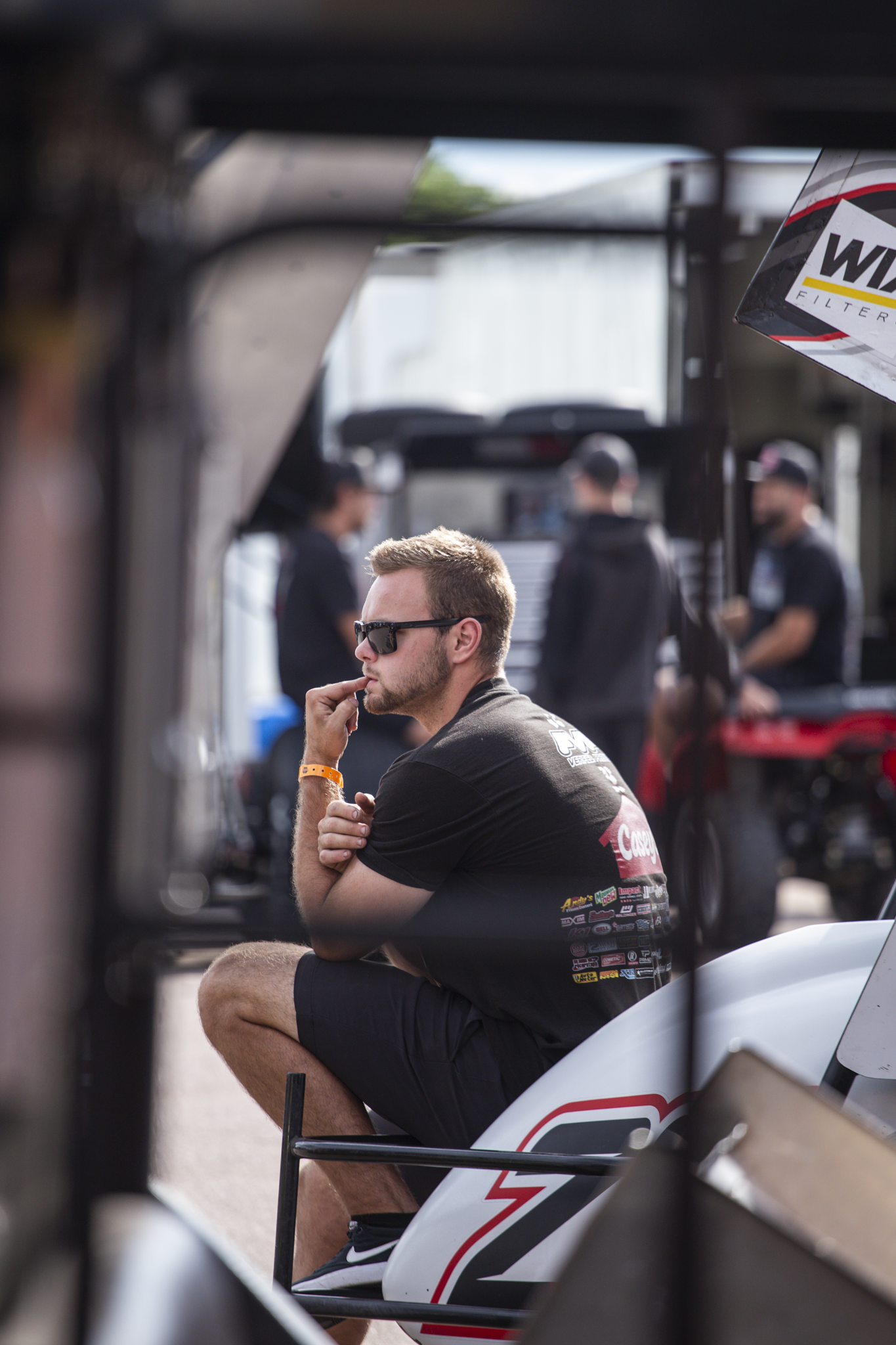 A young man with short light brown hair, wearing sunglasses, a black t-shirt with multiple sponsor logos, and black shorts, sitting with one knee up and resting his chin on his hand, in a pit area at a racing event. Several blurred people are standing in the background.
