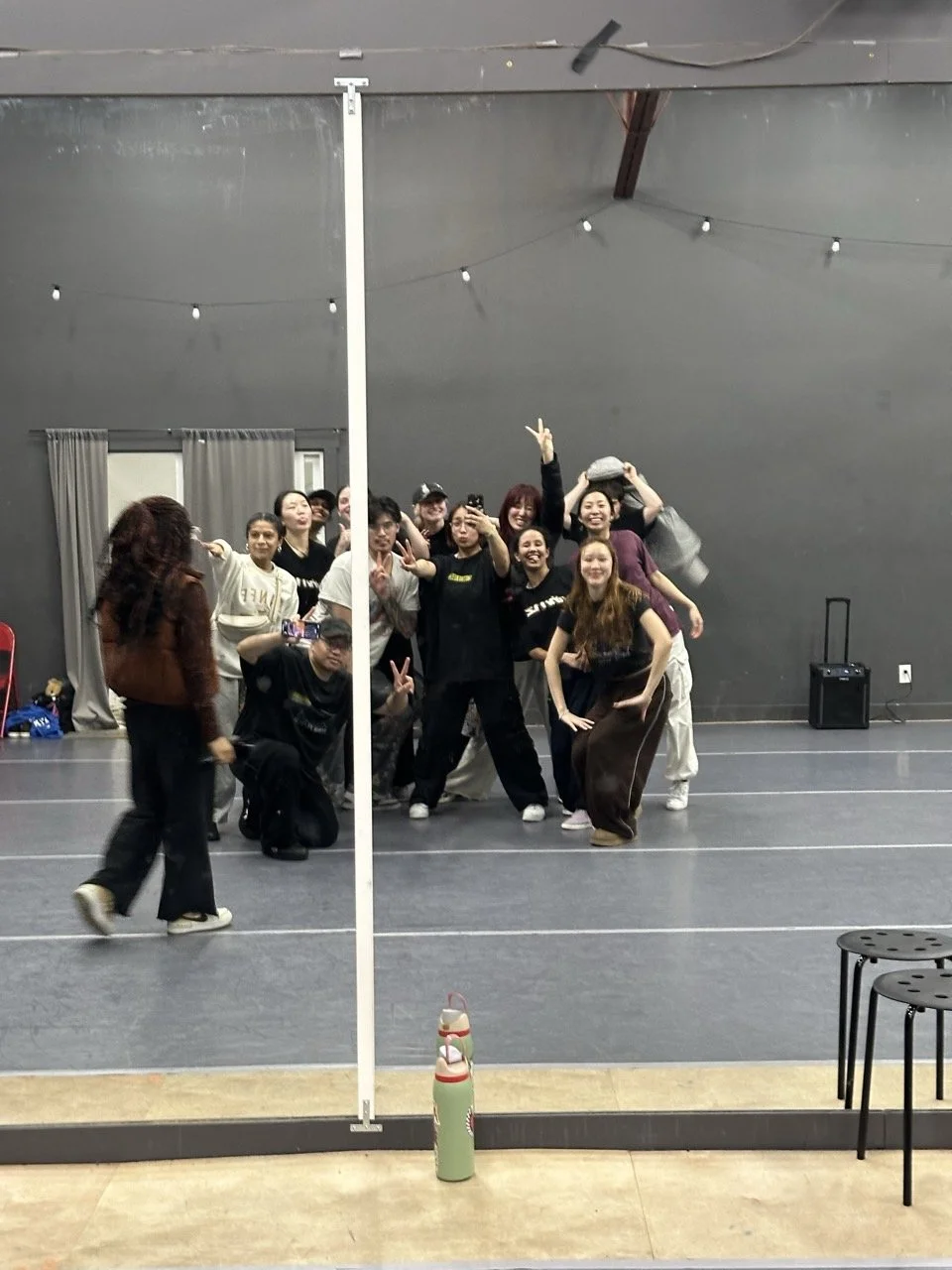A group of people posing for a photo in a dance studio, with some making peace signs, and one person holding a gray hat on their head. There are chairs and a water bottle in the foreground.