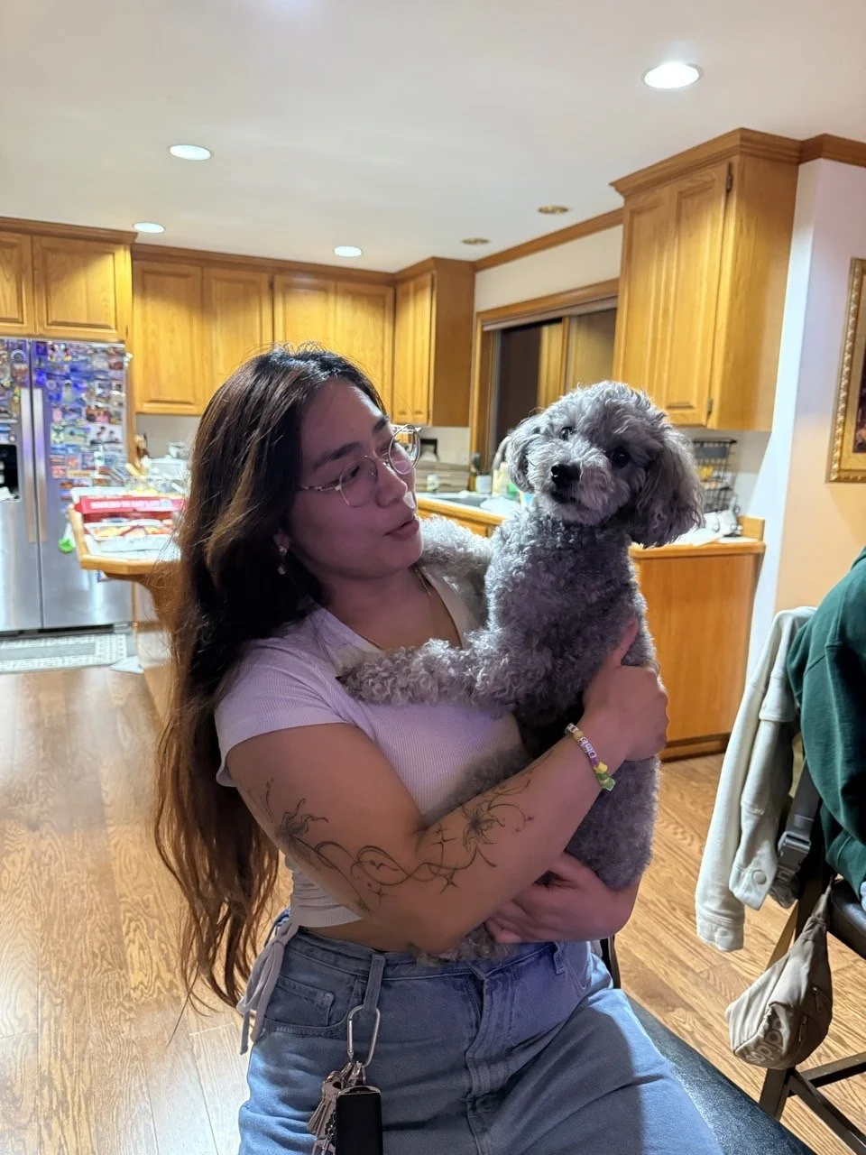A woman with long hair, glasses, and tattoos on her arm, holding a gray poodle in a kitchen with wooden cabinets.