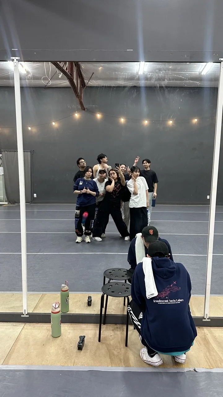 A group of eight people is posing for a photo in a dance studio, with two people crouching in front. The photo is being taken through a mirror, and there are string lights hanging from the ceiling. Two water bottles and small objects are on the floor in front of the mirror. The studio has a gray wall and a wooden floor.