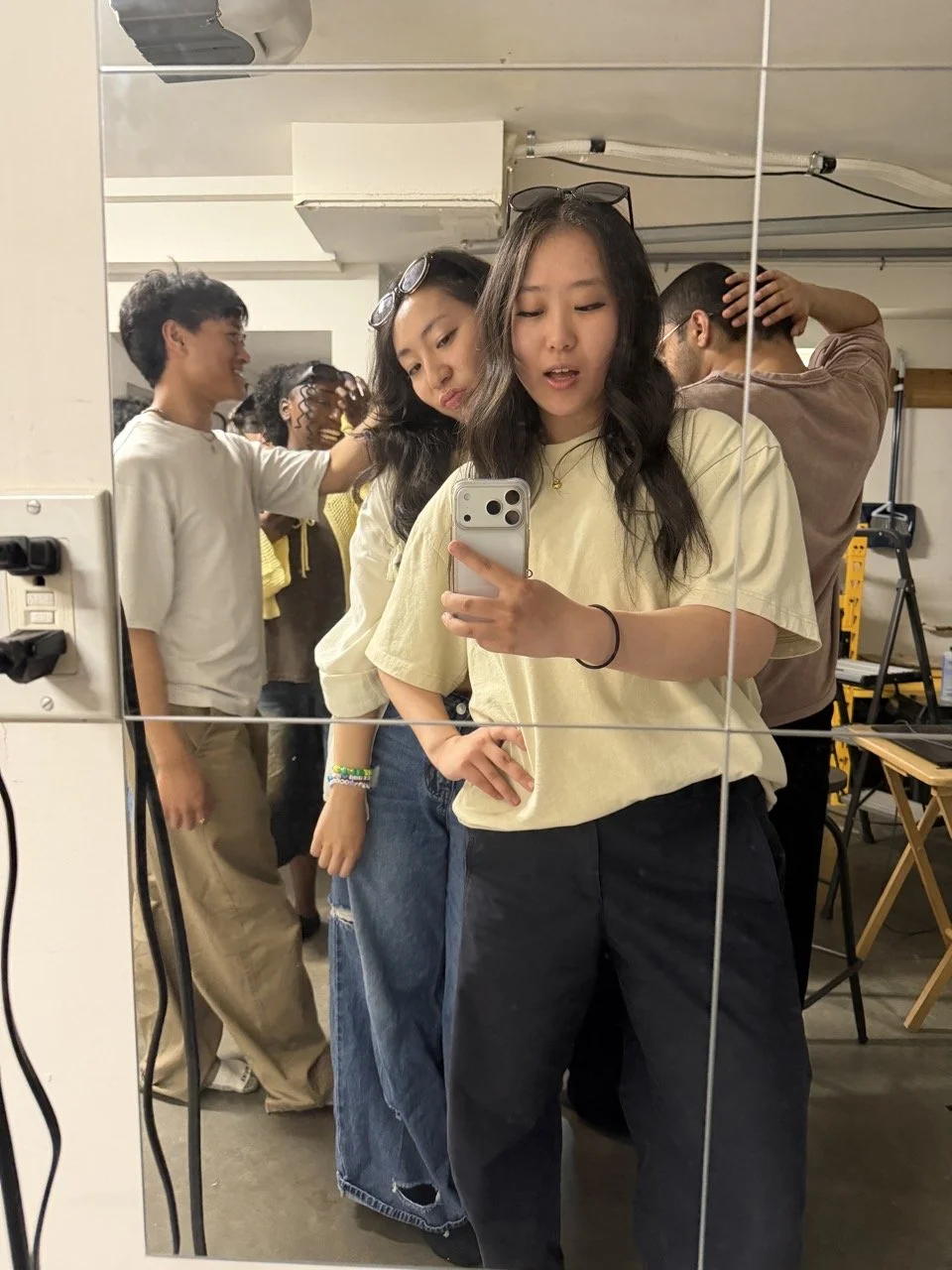 Group of diverse young people taking a selfie in front of a mirror, with some posing and others adjusting their hair, in a room with equipment and chairs.