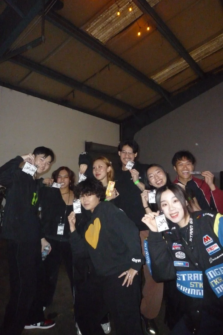 Group of nine young people in casual clothing smiling and holding up event passes indoors with a concrete ceiling and industrial lighting.