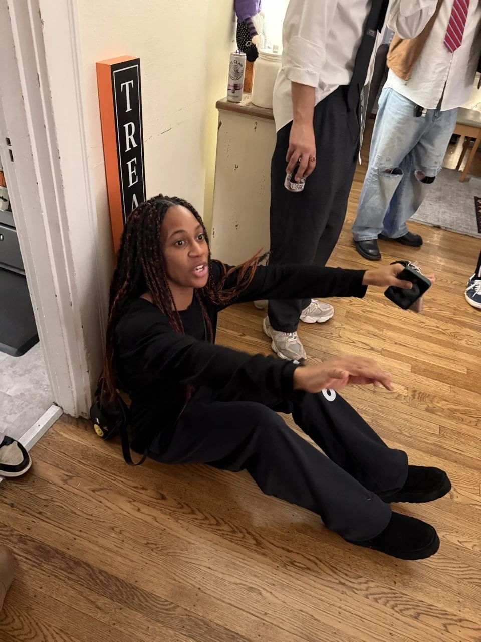 A woman with long braided hair sitting on the floor against a wall, holding a camera and talking. She is engaged in conversation with two men standing nearby in a room with wooden floors.