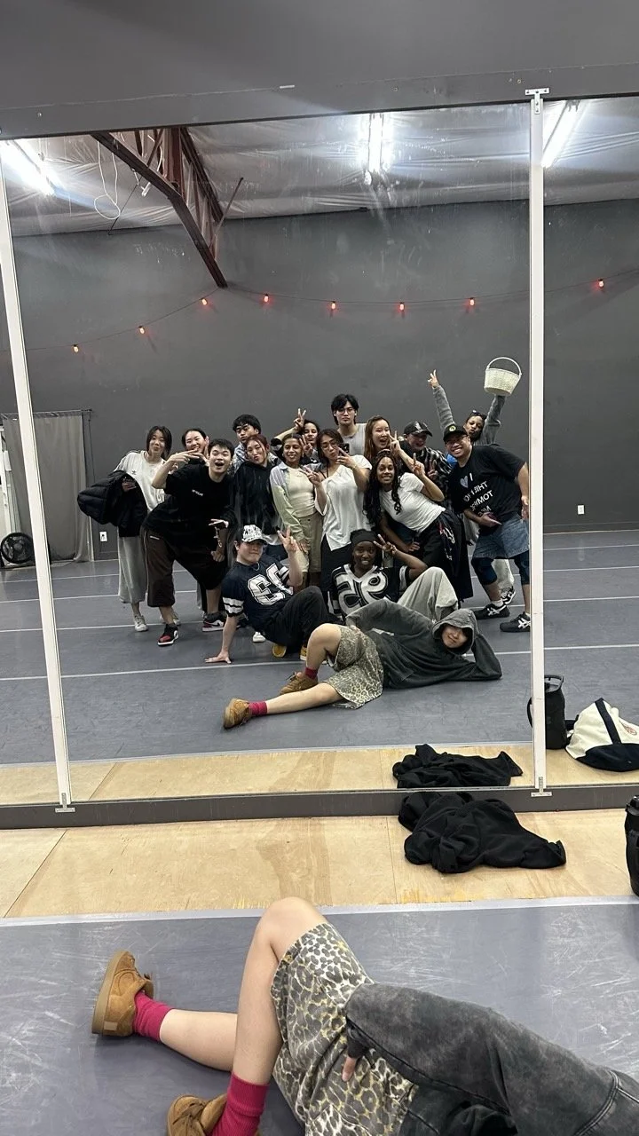 Group of young people posing and making peace signs in front of a mirror in a dance studio, with one person lying on the floor in front of the mirror.