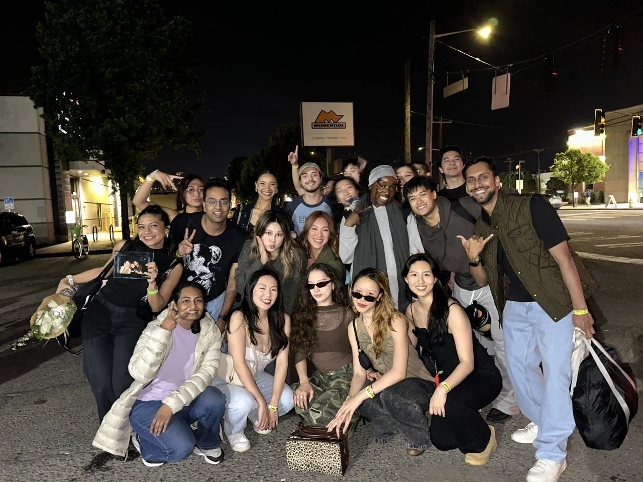 A group of 17 diverse people posing together on a city street at night, some smiling, making peace signs, and enjoying each other's company.