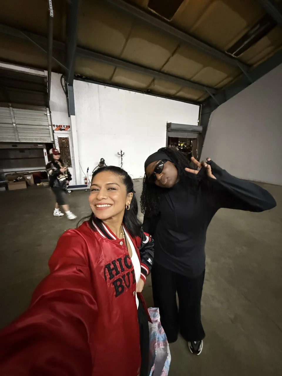 Two women taking a selfie inside a warehouse. One is wearing a red jacket that says 'Chic But' and is smiling at the camera. The other is wearing black sunglasses and a black outfit, making a peace sign. There is a person in the background walking by