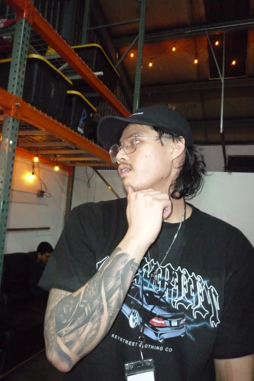 A young man with tattoos on his arm, glasses, and a black cap, touching his chin in a warehouse or industrial space with shelves and warm lighting.