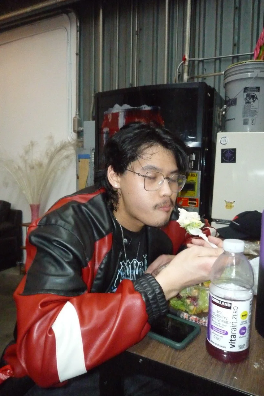 A man with glasses and a goatee eating a salad with a fork, sitting at a table with a bag of Salad, a water bottle, and a smartphone, in an industrial-style setting.