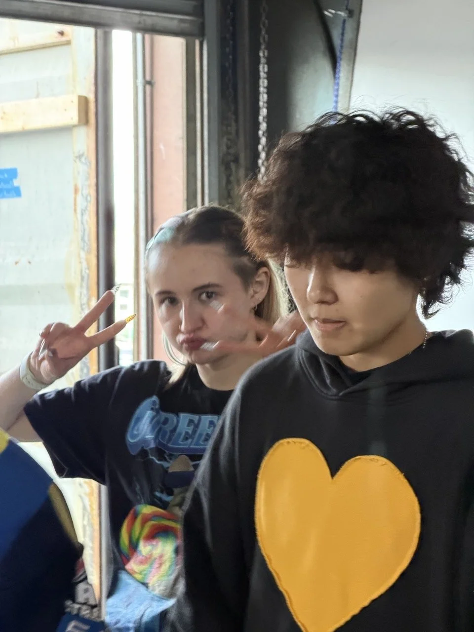 Two young people standing inside near a door, with the girl making a peace sign and the boy looking down.