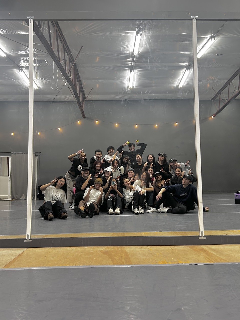 Group of young people posing and smiling for a photo in a dance studio or rehearsal space, with some making peace signs and others holding gym or dance equipment.