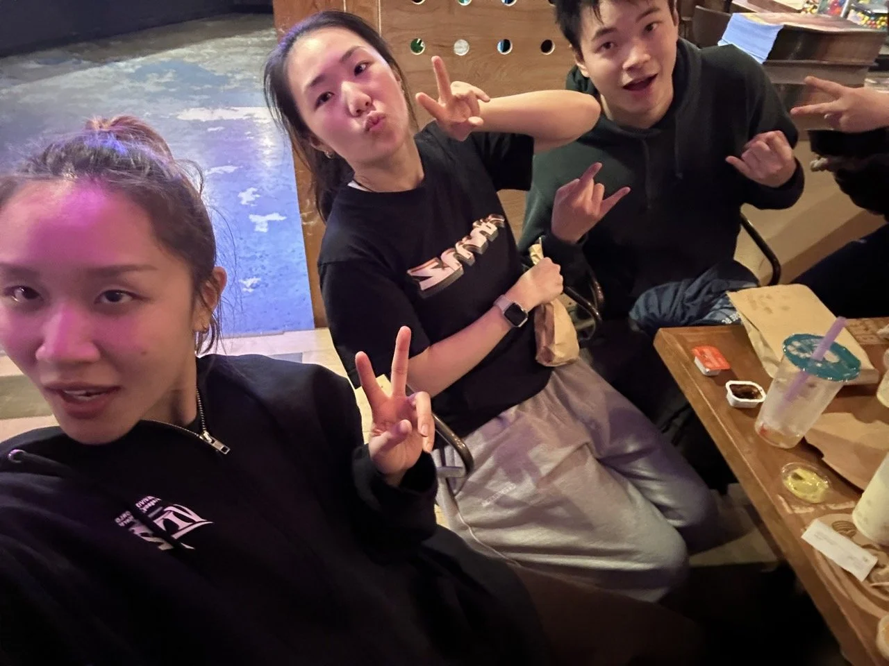 Three young people sitting at a table in a restaurant or cafe, making peace signs and posing for the camera, with drinks and snacks on the table.