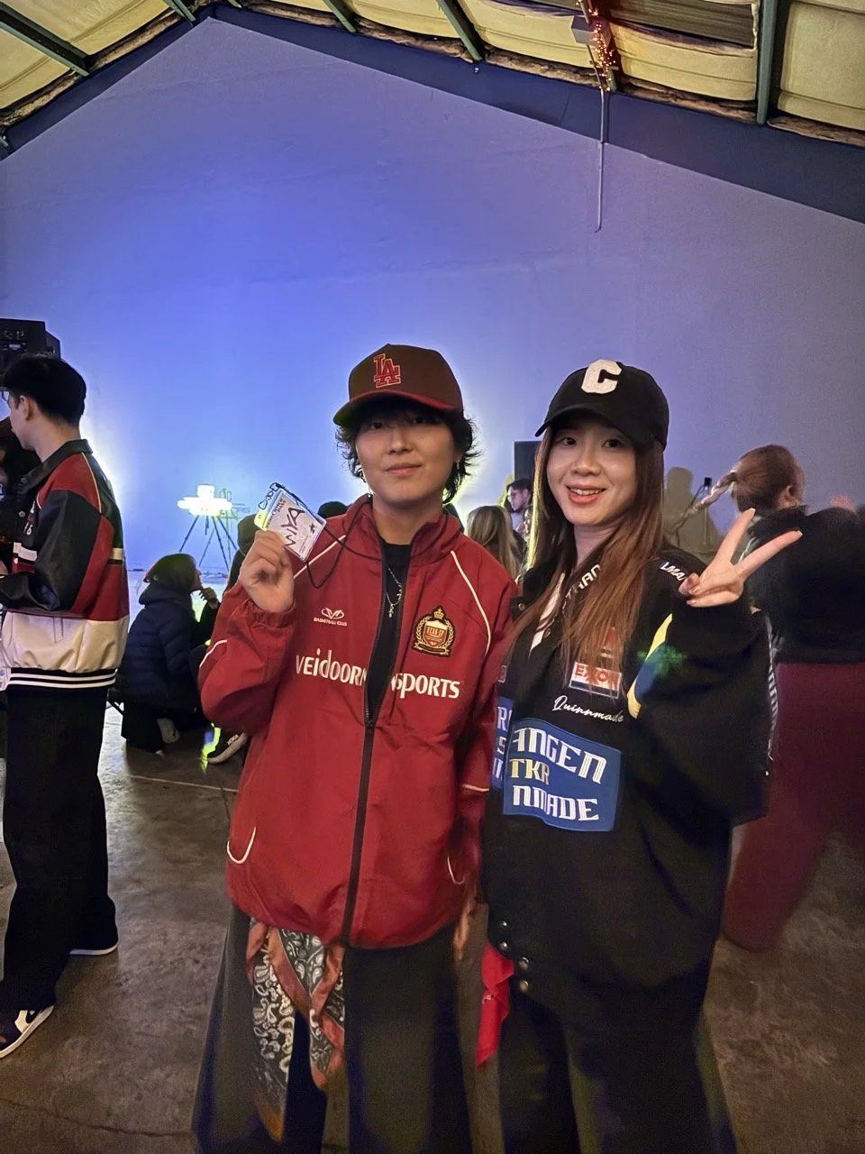 Two young women posing for a photo at an indoor event. One is wearing a red jacket and a red LA baseball cap, holding a lanyard. The other is wearing a black jacket with various patches and making a peace sign. Several people are in the background, a