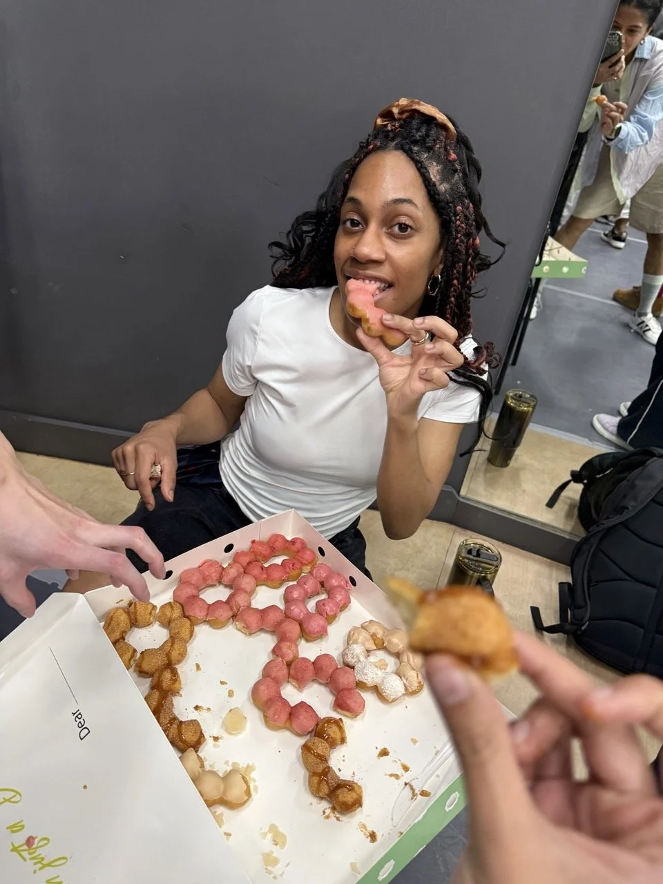 A woman with braided hair licking a donut, sitting in front of a box of colorful donuts with pink, white, and brown frosting, as people in the background look on.