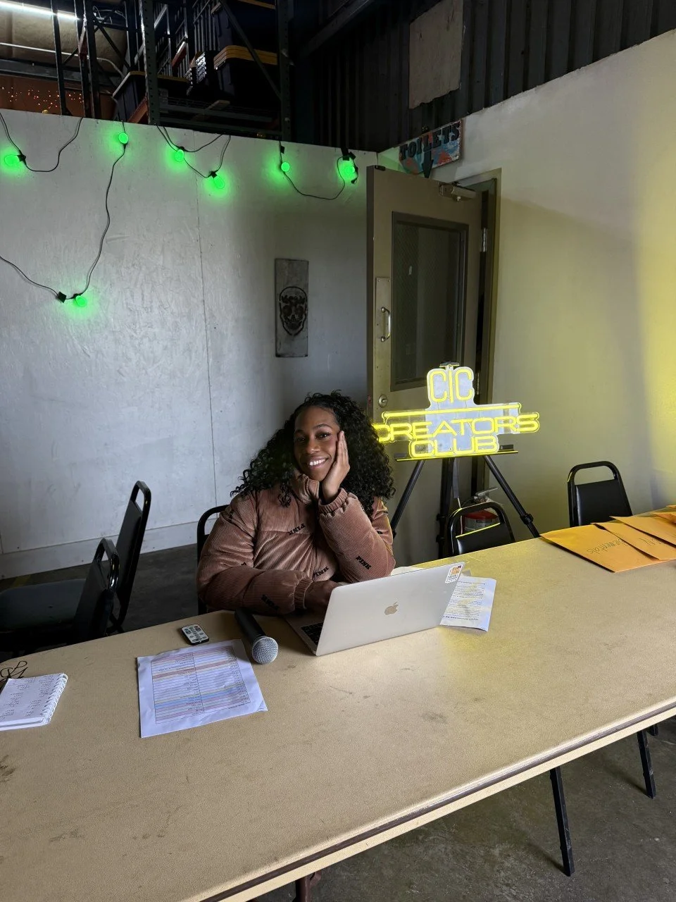 A woman sits at a yellow conference table with a laptop, papers, and a microphone in front of her, smiling with her hand on her cheek. Behind her, there is a neon sign that says 'CIC CREATORS CLUB' and a display of green string lights on the wall.