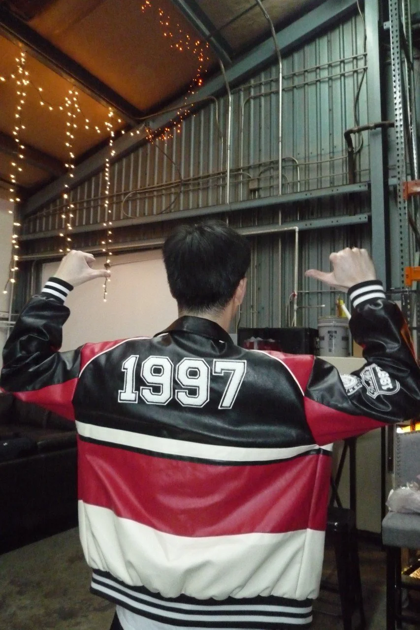 Person wearing a black, red, and white varsity jacket with '1997' on the back, standing in a warehouse-like space decorated with string lights, facing away from the camera, with arms raised.