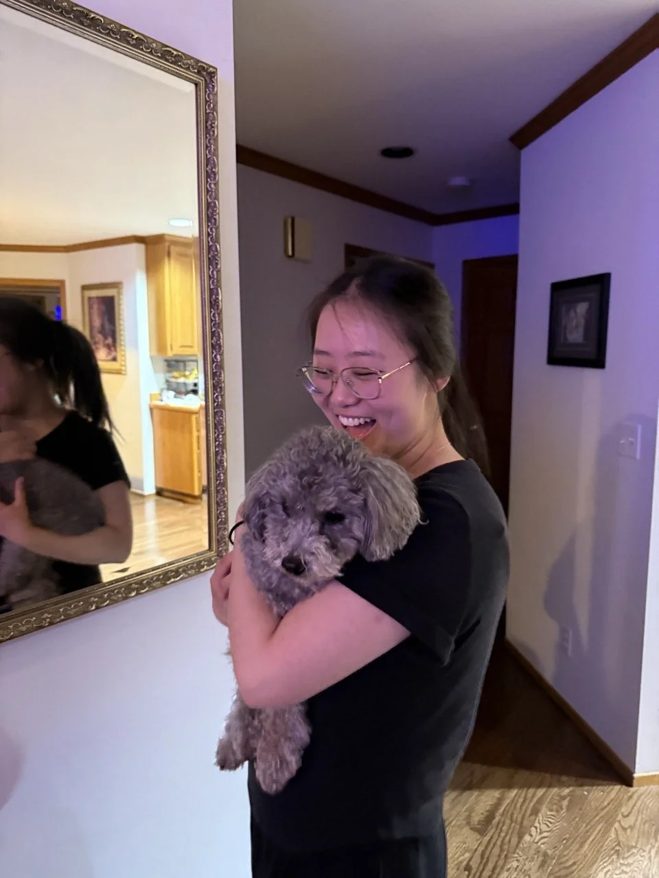 A woman with glasses is smiling while holding a fluffy gray puppy in her arms inside a house. The scene is warm and joyful.