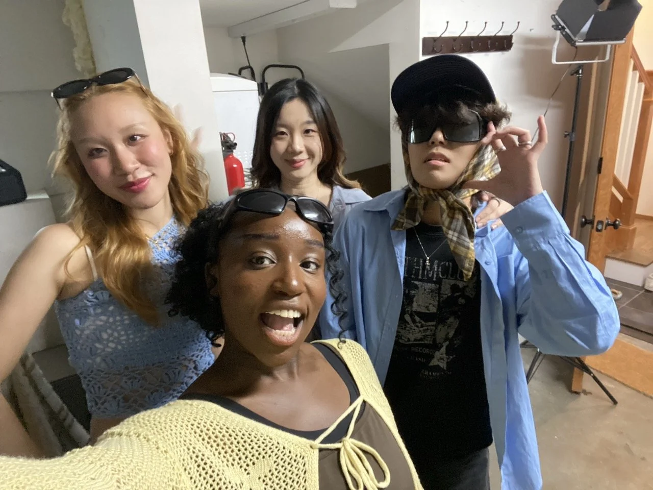 Four women posing indoors, some wearing sunglasses and accessories, with a background including a fire extinguisher, a coat rack, and studio equipment.