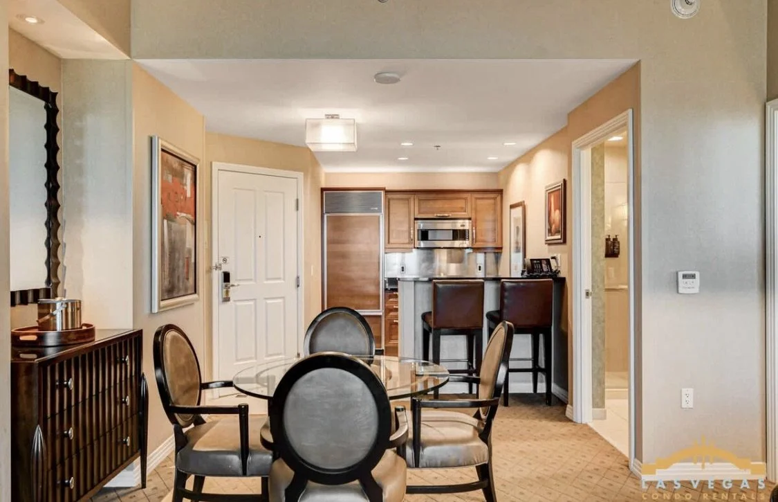 Dining area with a glass table and four upholstered chairs, adjacent to a kitchen with wooden cabinets, stainless steel appliances, and a breakfast bar with two chairs.