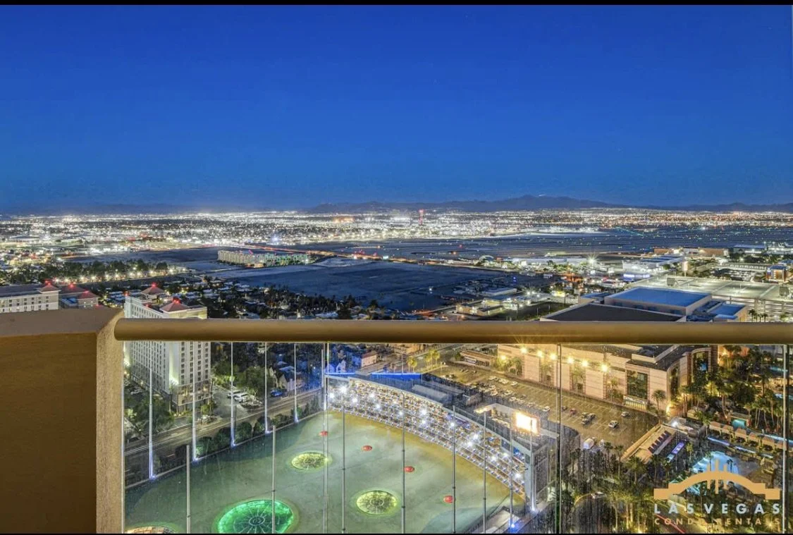 Nighttime view from the balcony overlooking Las Vegas with city lights, mountains in the distance, and Top Golf below with colorful lighting.