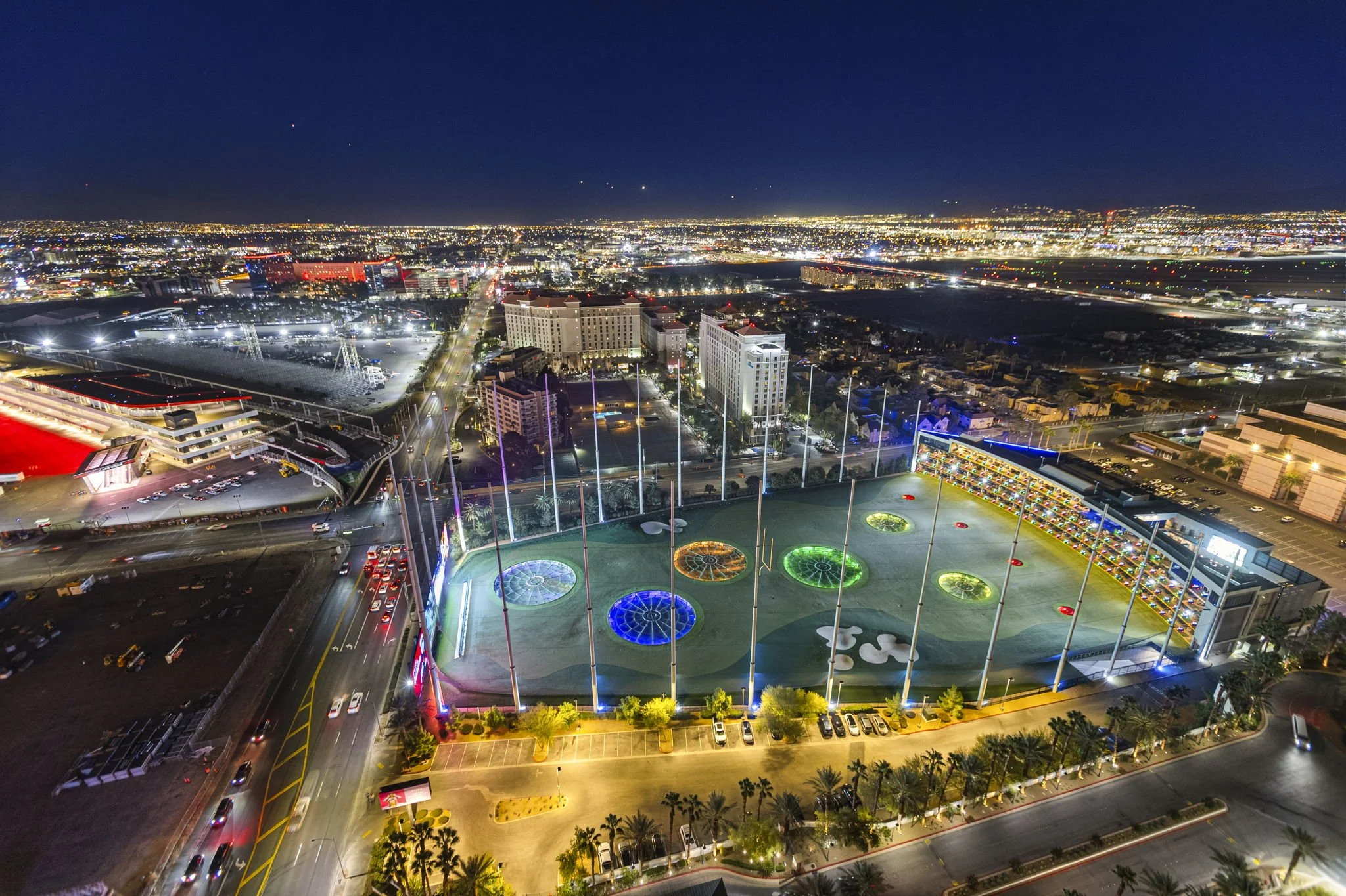 Night aerial view of a city with illuminated buildings, streets, and a brightly lit parking lot. Foreground features a rooftop golf driving range with colorful illuminated circular targets.