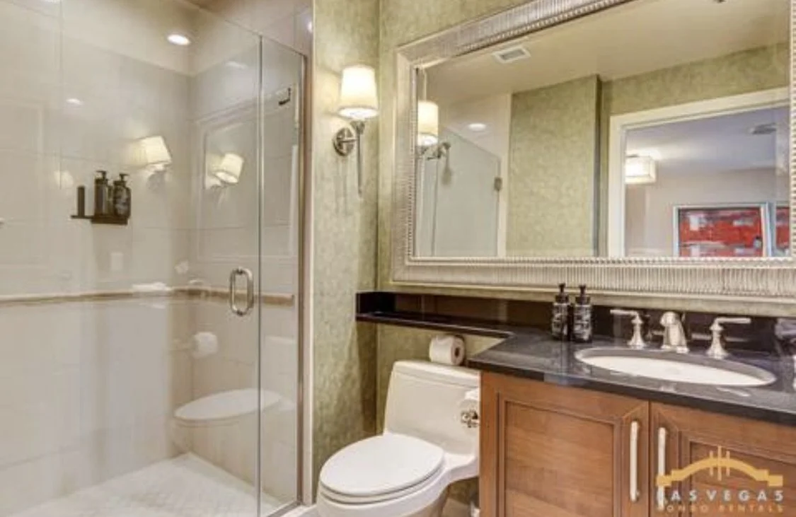 Guest bathroom with a glass-enclosed shower, a toilet, dark countertop with a sink, large framed mirror, and wall-mounted light fixtures.
