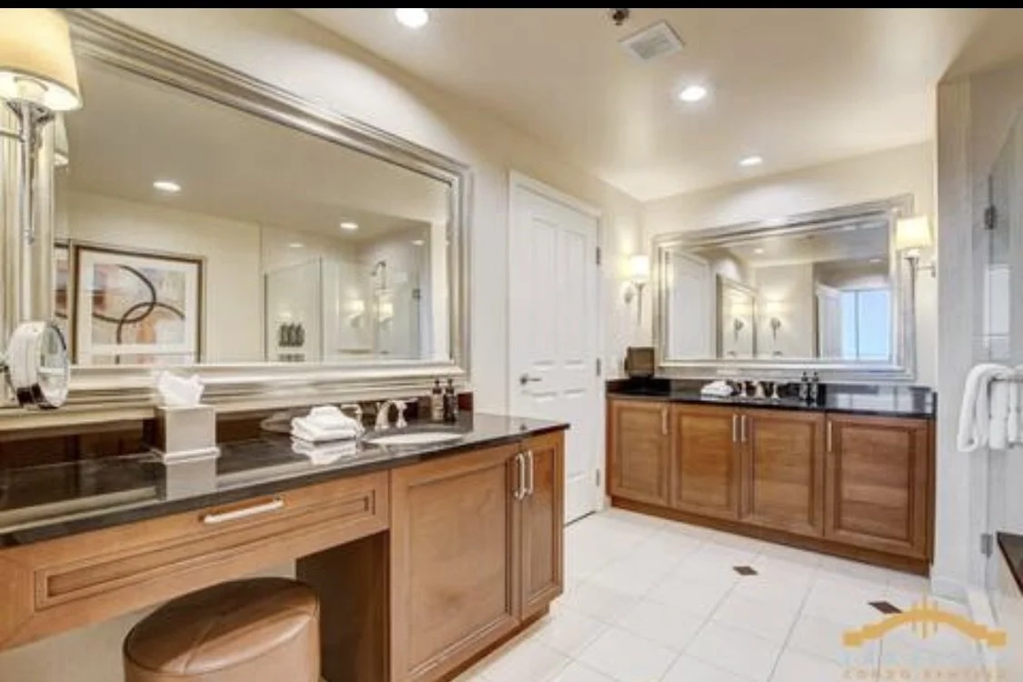A luxurious bathroom with two wooden vanities, large wall mirrors, a stool, and beige tiled floor.