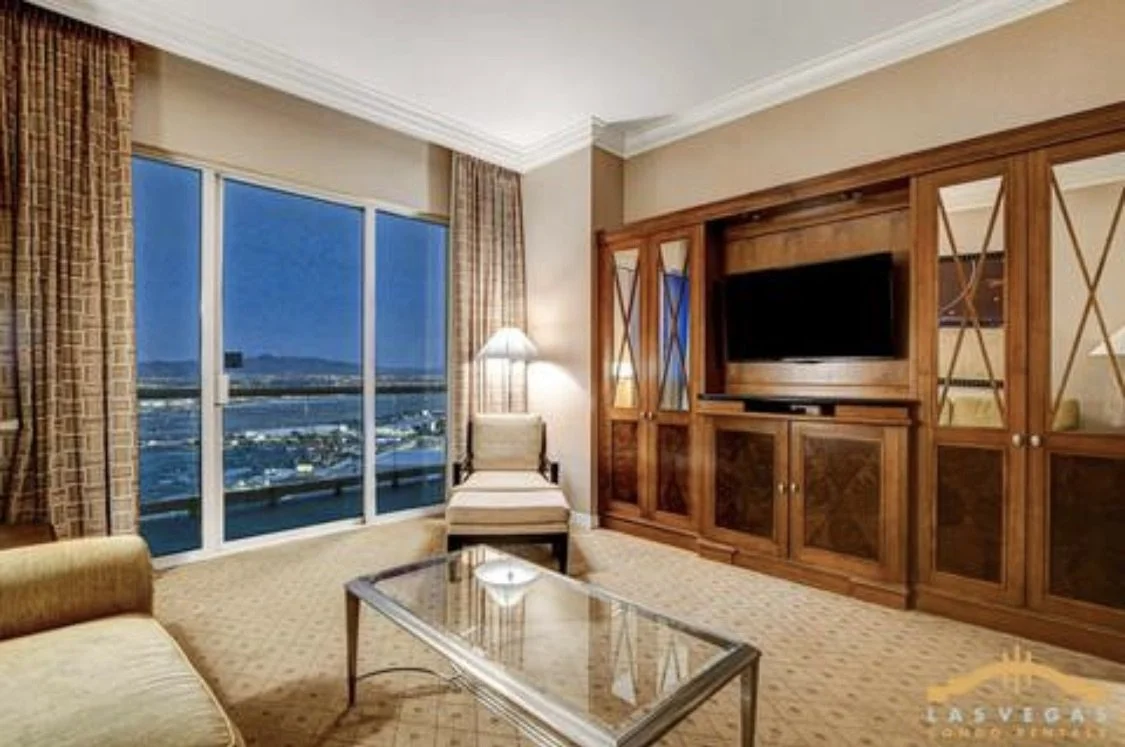 Penthouse suite living room with 10 foot ceiling, large windows and door to balcony showing city view.  Entertainment center with television, armchair by window, glass coffee table, and a sofabed in the foreground.