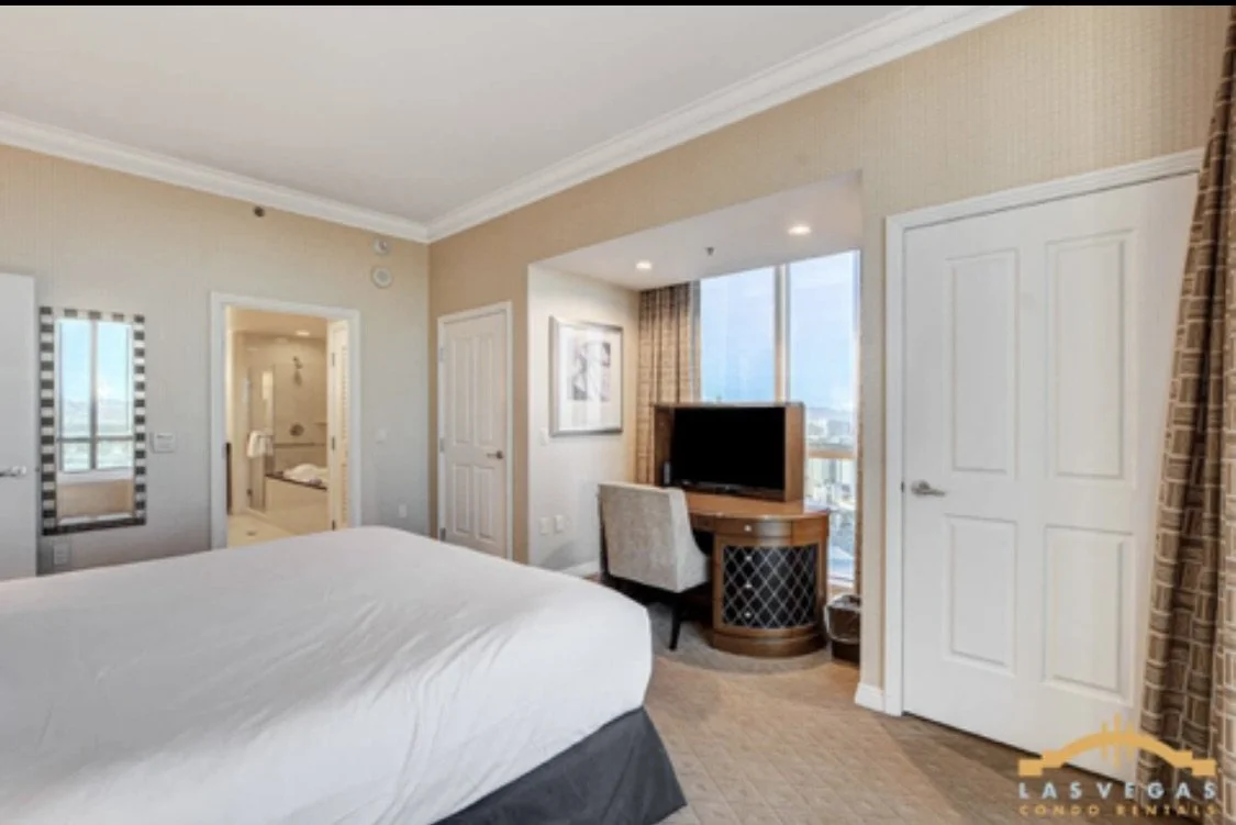 Suite bedroom with a king bed, TV and wooden desk, window with city view, master bathroom entrance, and curtains.