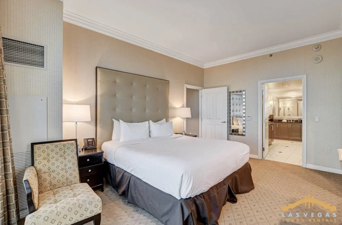 The bedroom with a large king bed, two nightstands with lamps, a chair, a mirror, and a doorway leading to the master bathroom.