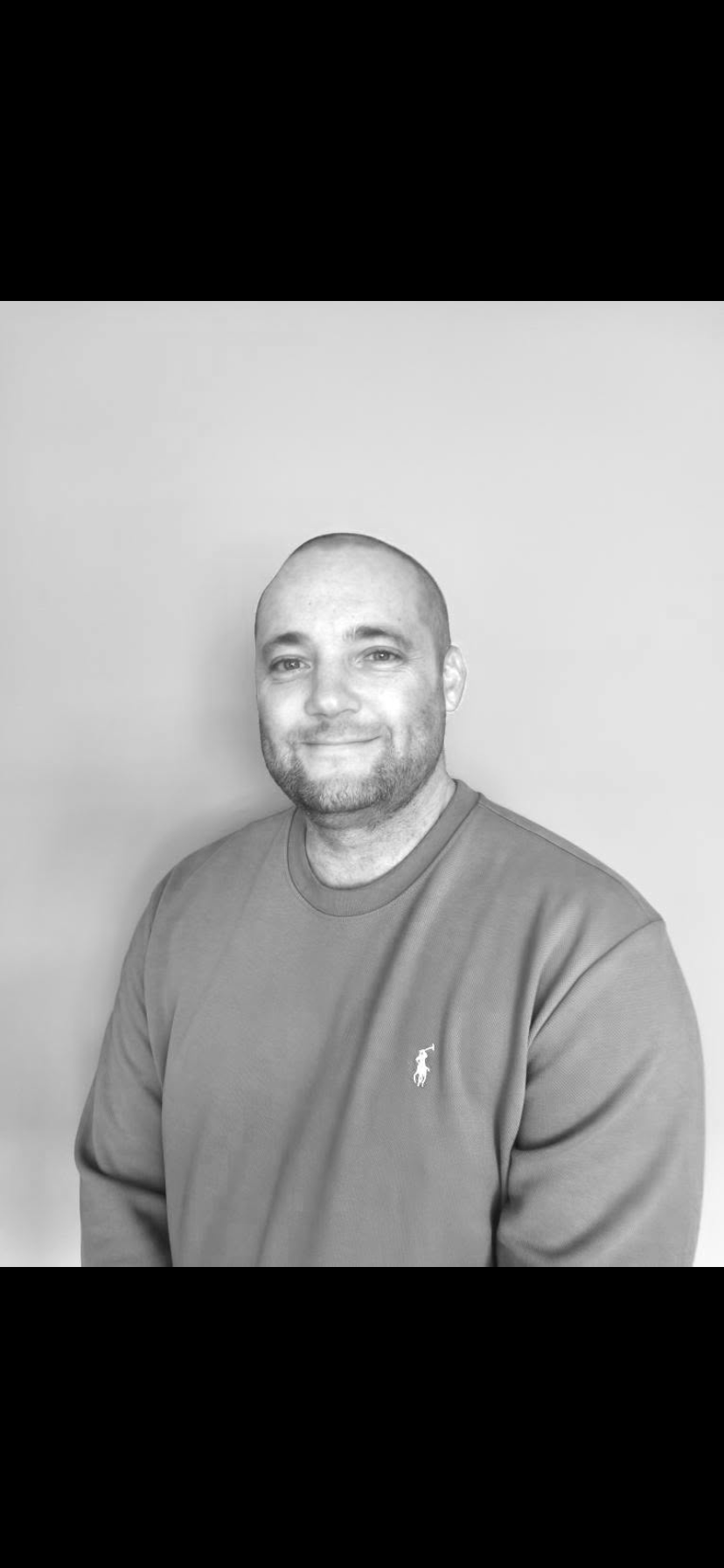 Black and white photo of a man with a beard smiling, wearing a sweatshirt with a small logo of a polo player on the chest, standing against a plain wall.