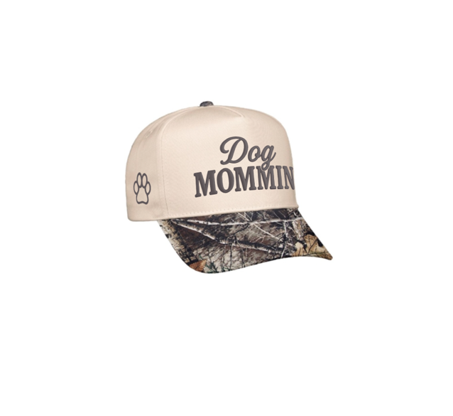 Beige baseball cap with a camo pattern on the brim, featuring the words 'Dog MOMMIN' embroidered on the front and a small paw print on the side.