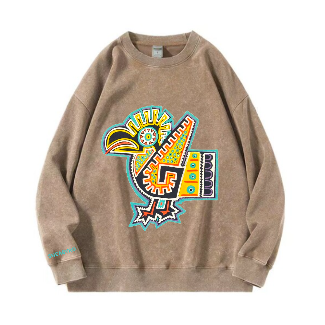 A beige sweatshirt featuring a colorful, abstract, tribal-style toucan illustration on the front, with the word "SHEABYRD" printed in blue on the lower sleeve.