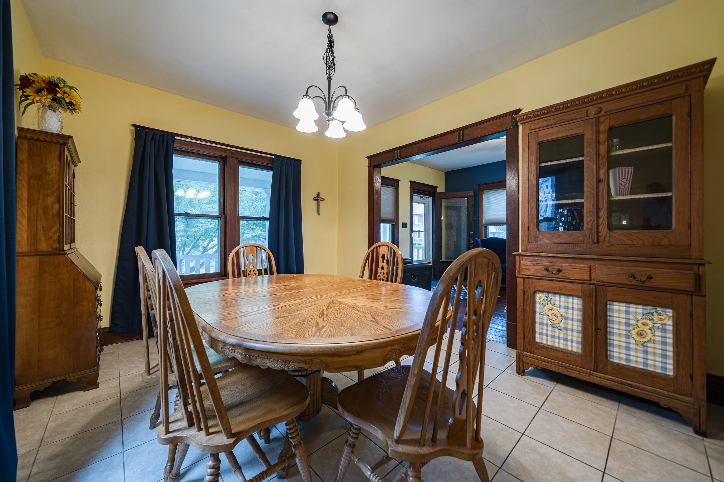Dining room with a wooden oval table and six matching chairs, yellow walls, a wooden hutch with flower pattern insets, windows with dark blue curtains, and a hanging ceiling light fixture.