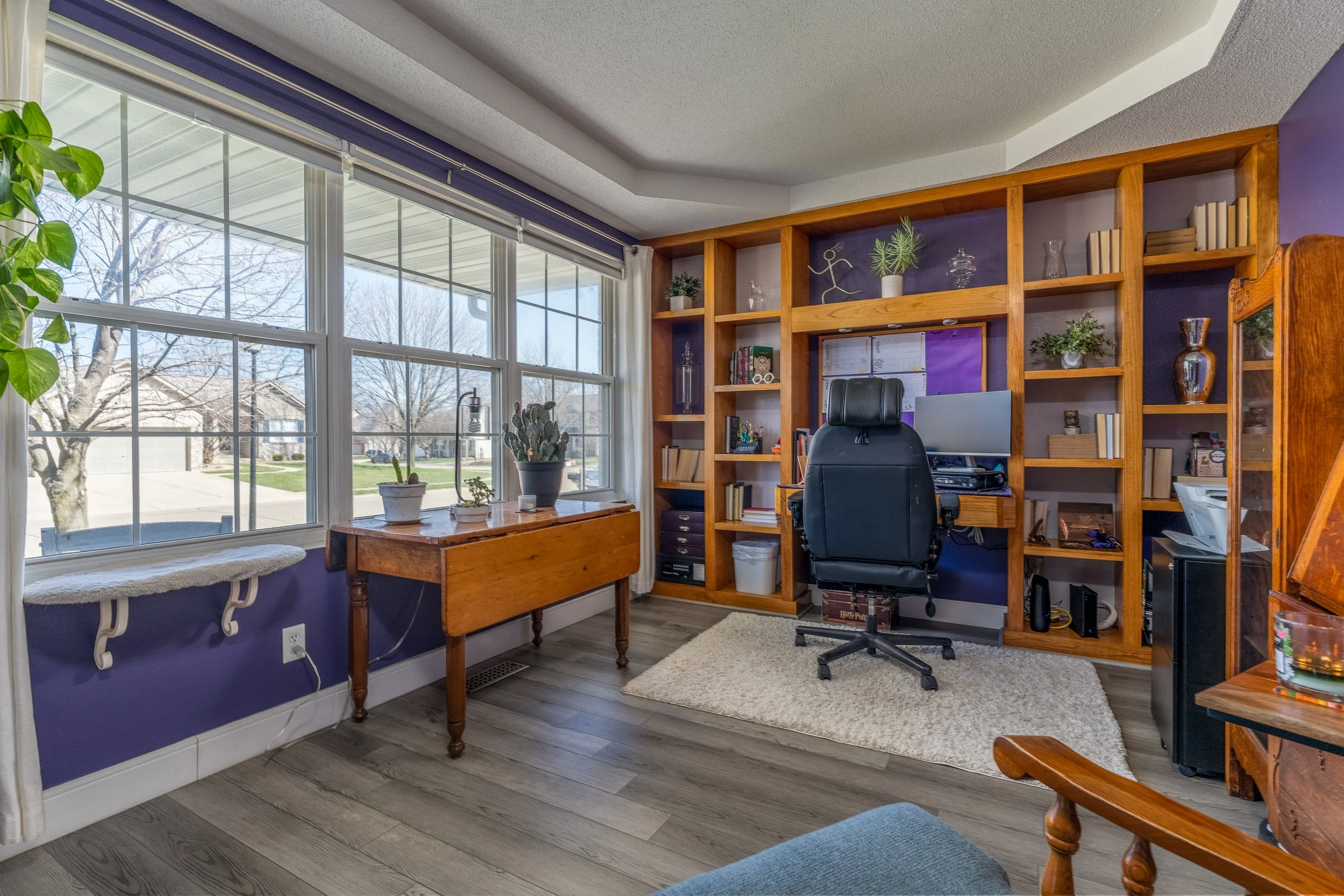 Home office with large window, purple walls, wooden bookshelf, black office chair, desk with computer, plants, and decorative items.