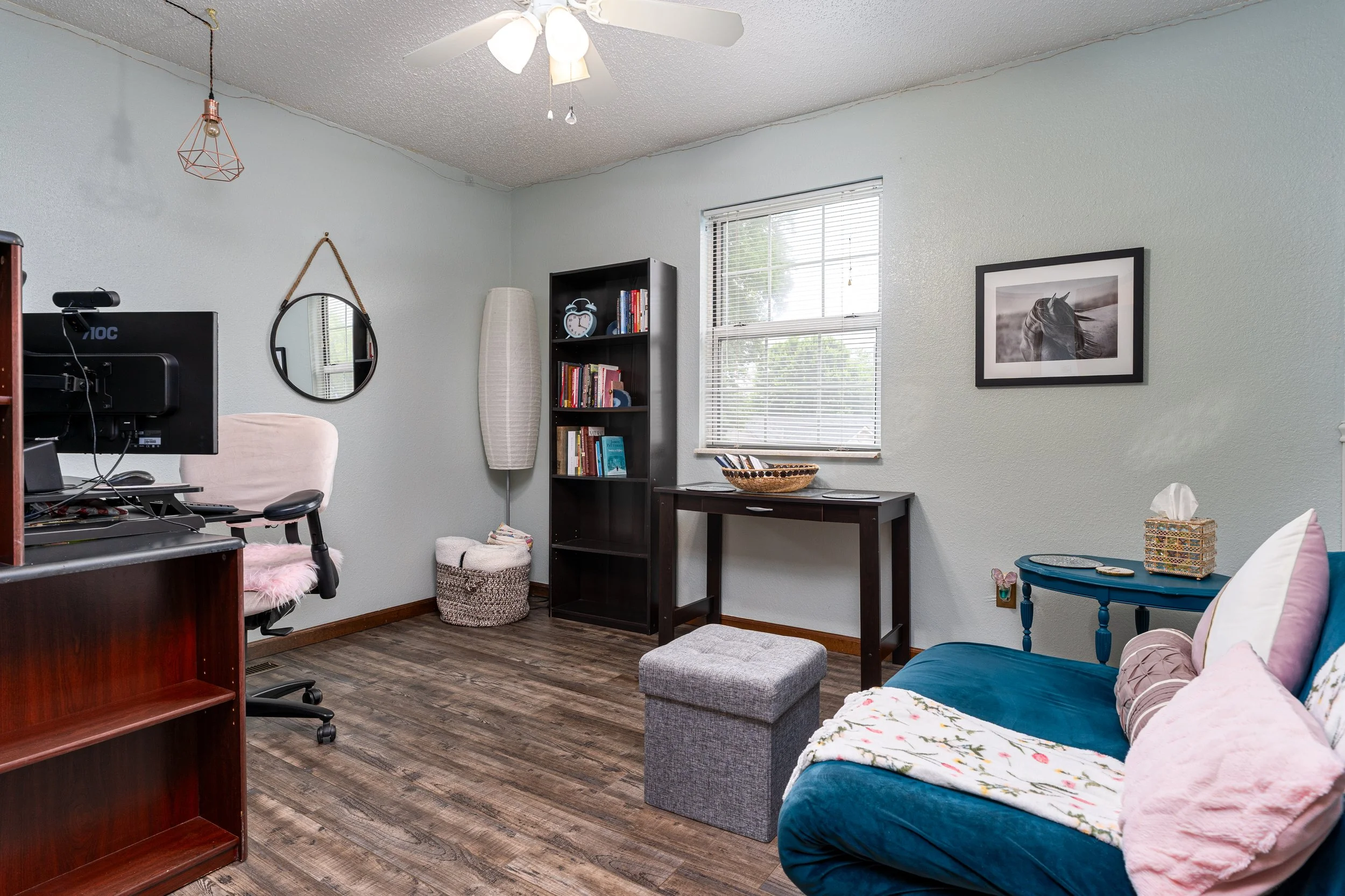Living room with a ceiling fan, a window with blinds, a blue sofa with pillows, a black bookshelf with books, a desk with a computer, a white floor lamp, and a wall picture of a horse.