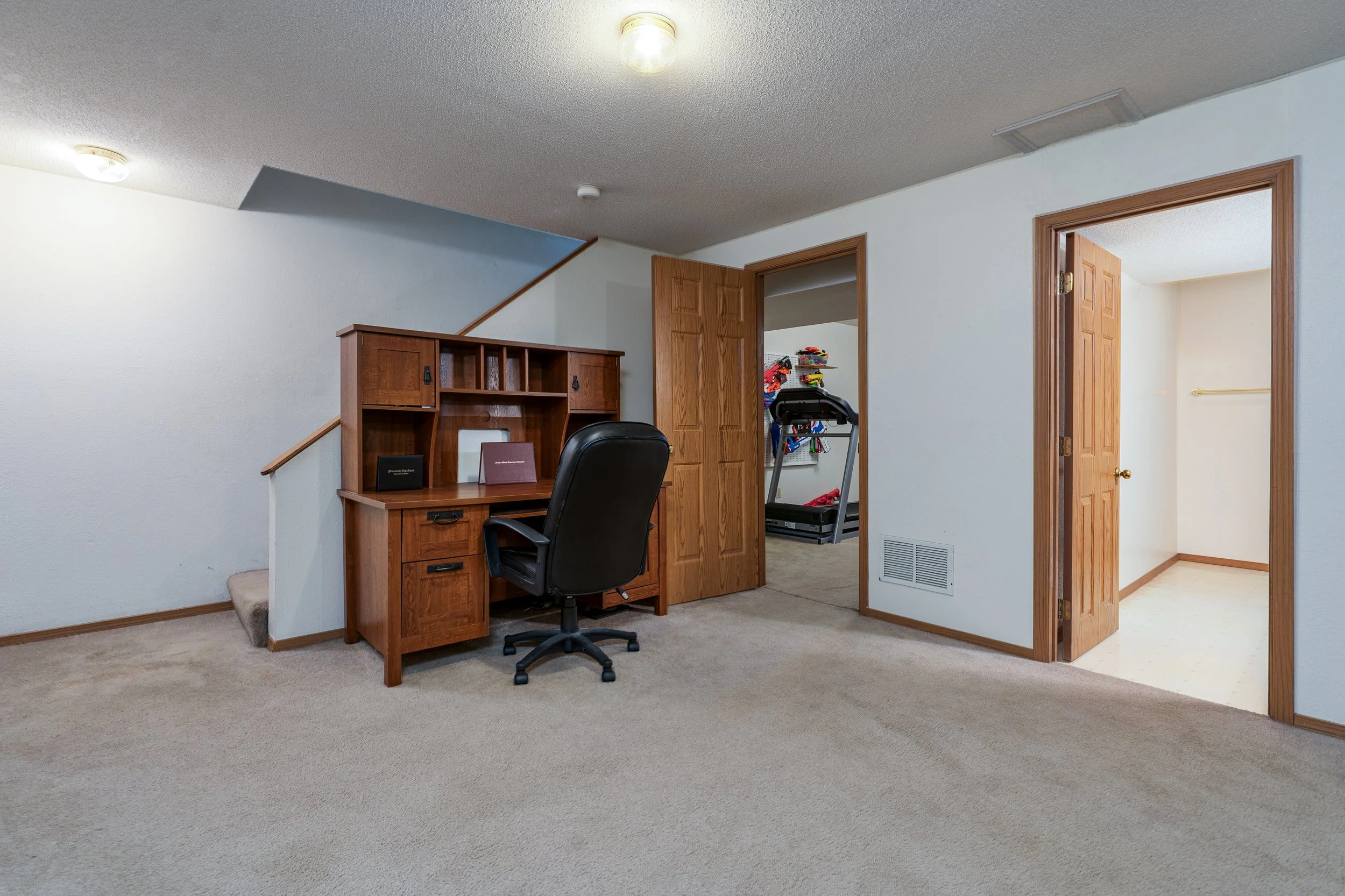 Home office with wooden desk and black office chair, closet with treadmill, carpeted floor, white walls, two open doors, ceiling lights.