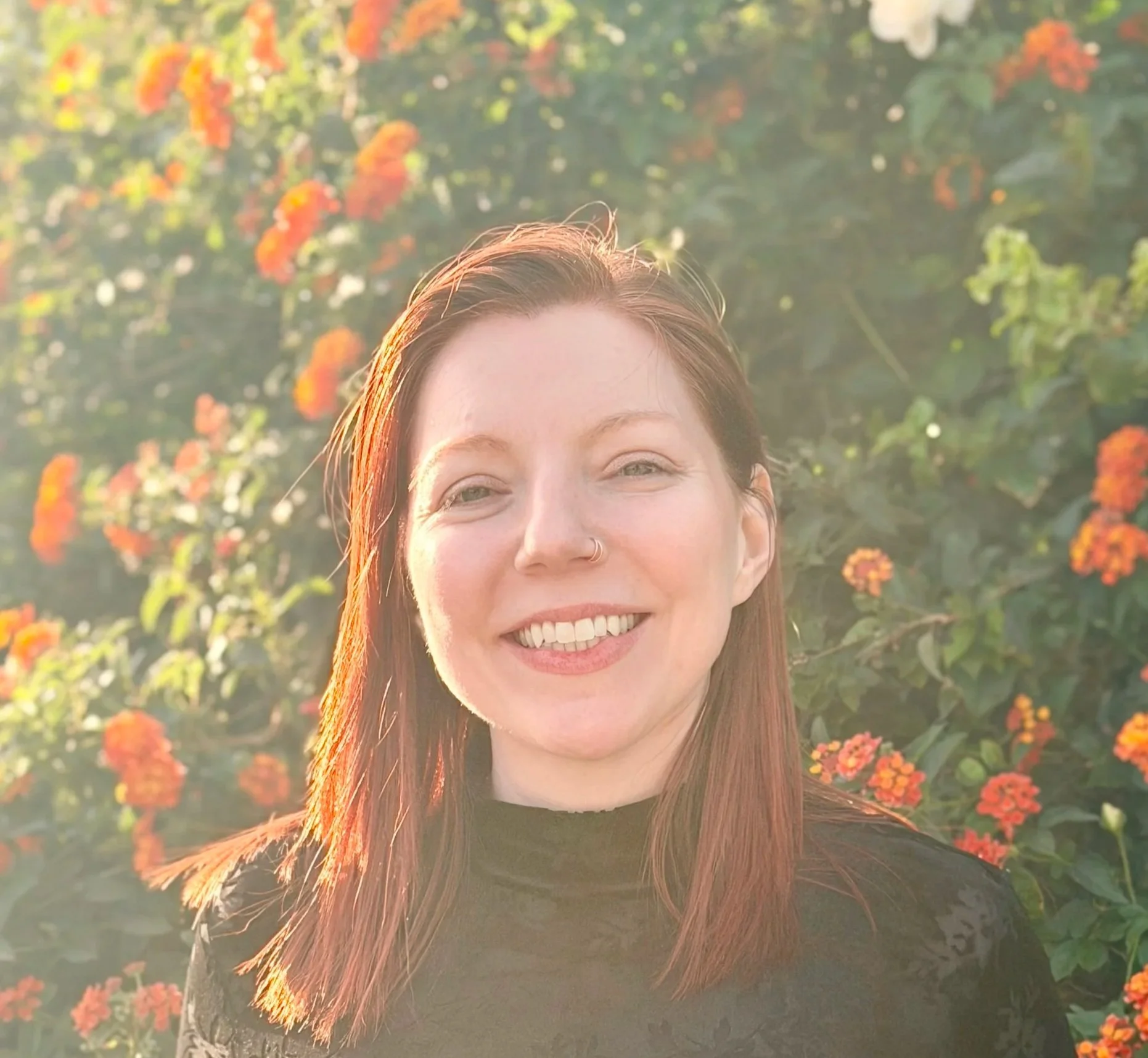 A woman with red hair smiling outdoors with orange flowers and green foliage in the background.
