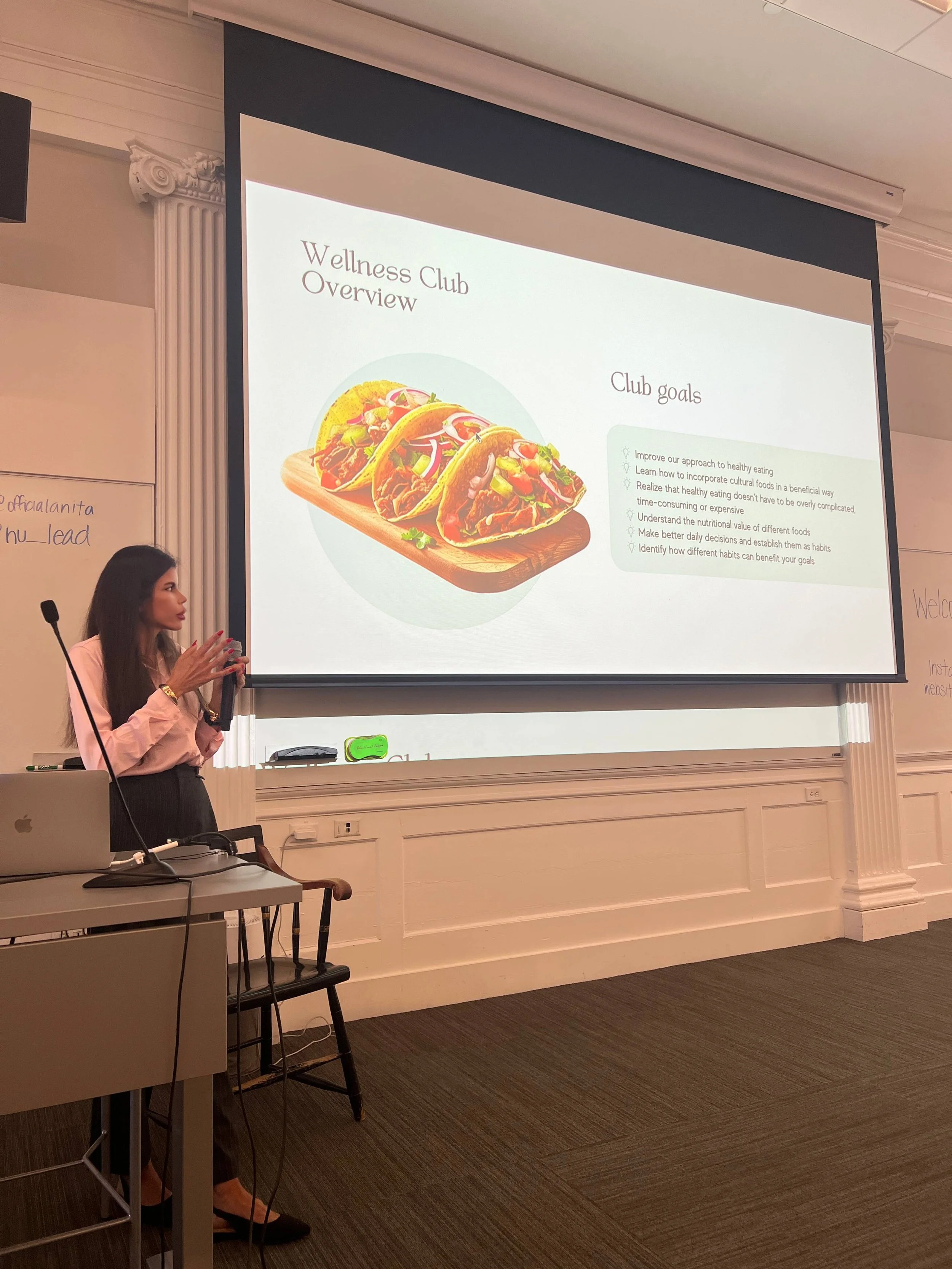 A woman giving a presentation in a conference room shows a slide titled 'Wellness Club Overview' with an image of tacos, and a list of club goals.