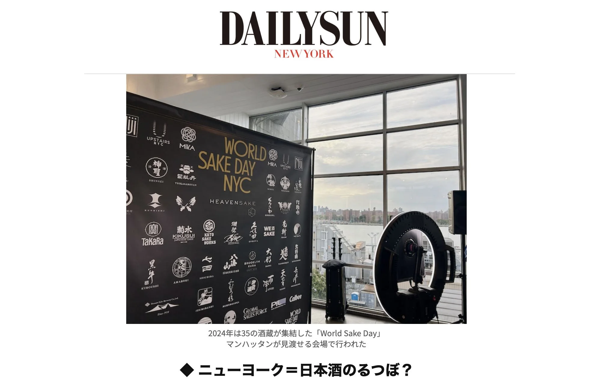 Indoor scene at World Sake Day NYC, with a large black banner displaying various sake brand logos and text, overlooking a city skyline through large windows, with some photography equipment visible.