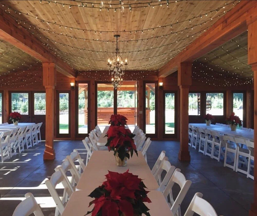 A decorated indoor event space with string lights, a chandelier, wooden beams, and tables adorned with white tablecloths and red poinsettia centerpieces.