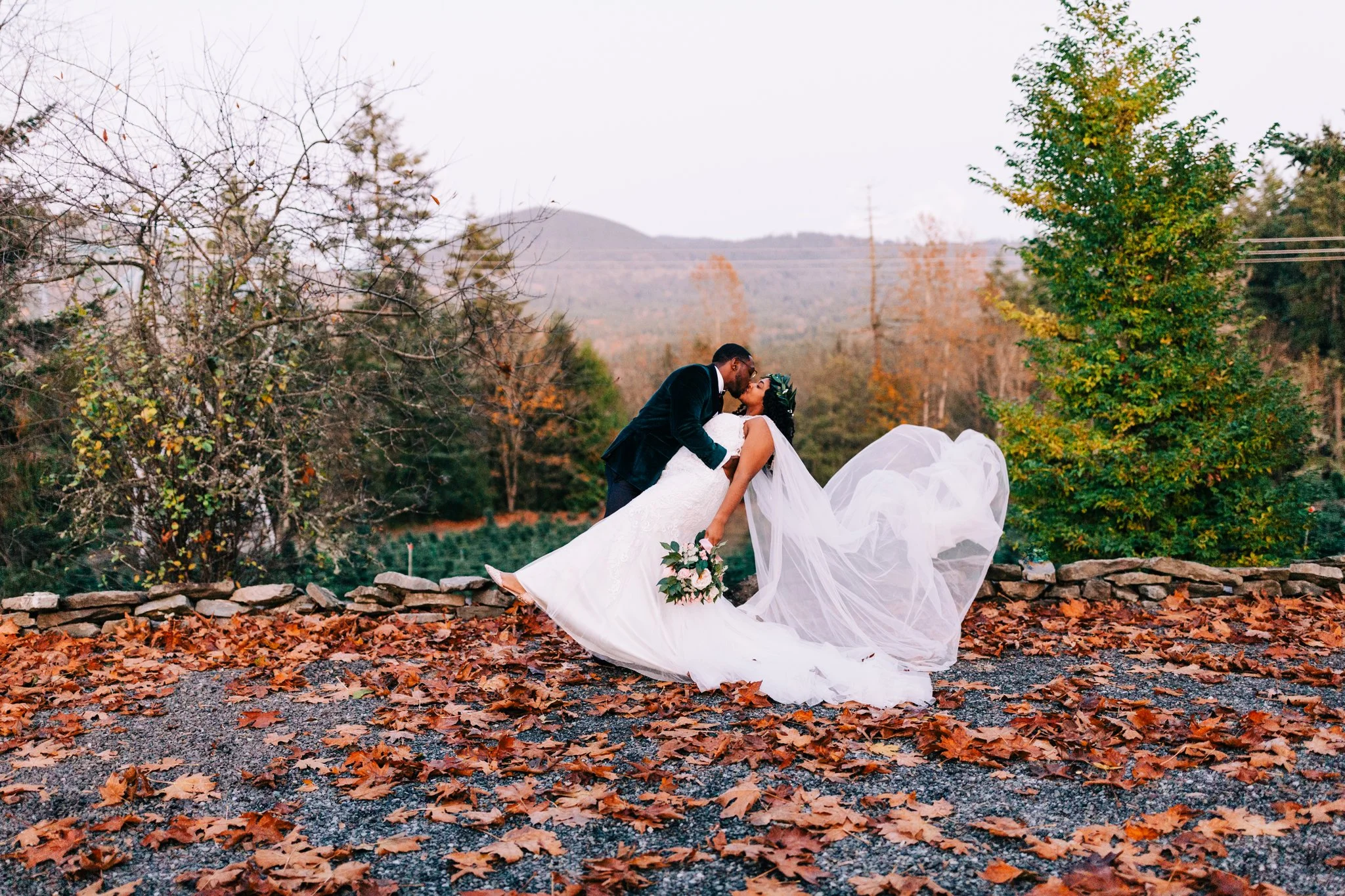 A newlywed couple sharing a kiss outdoors during autumn, with fallen leaves on the ground, trees with changing leaves, and a mountain in the background.