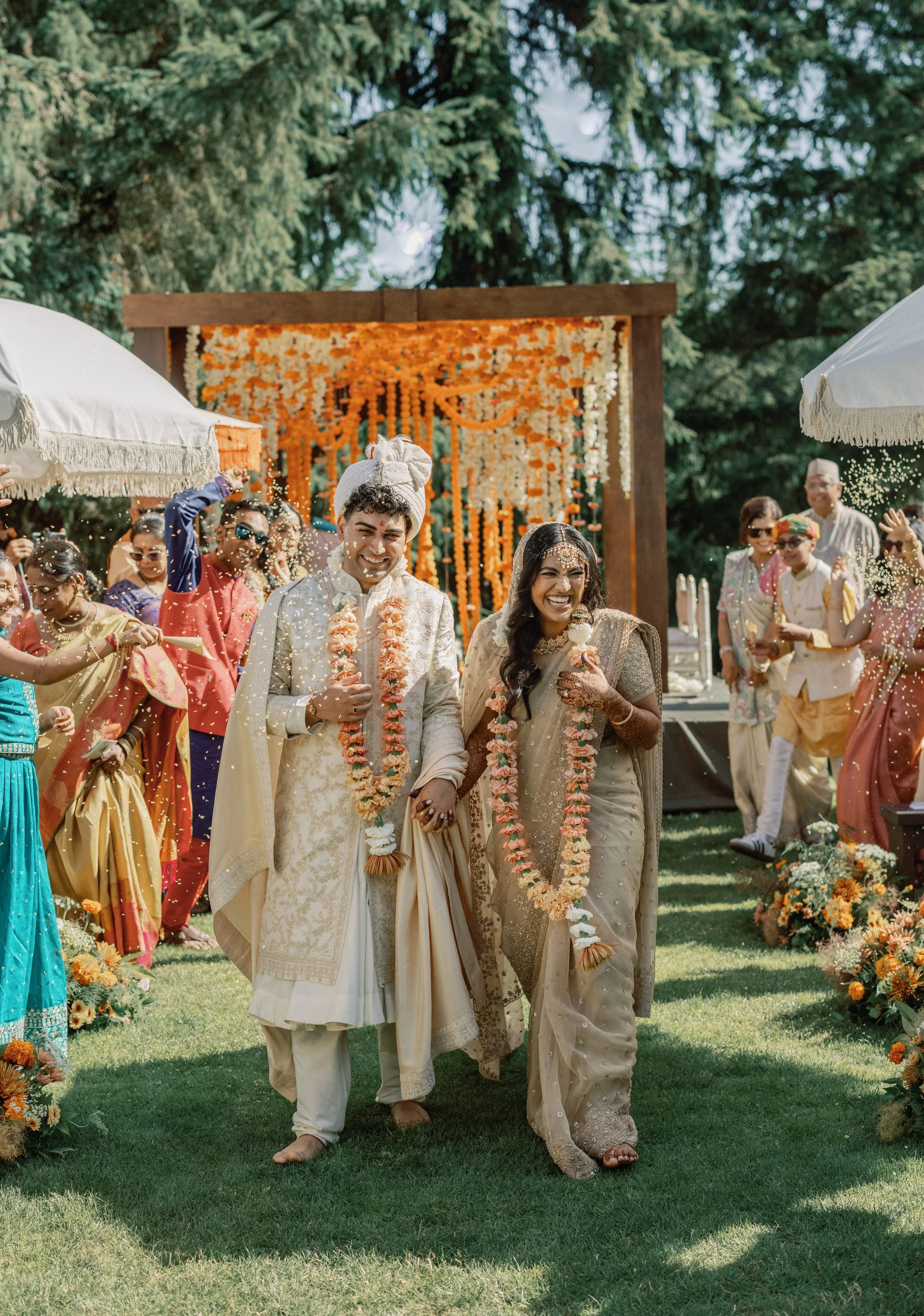 A newlywed Indian couple walking hand-in-hand through a garden, smiling as they are showered with flower petals during their wedding celebration, with guests in colorful traditional attire surrounding them.