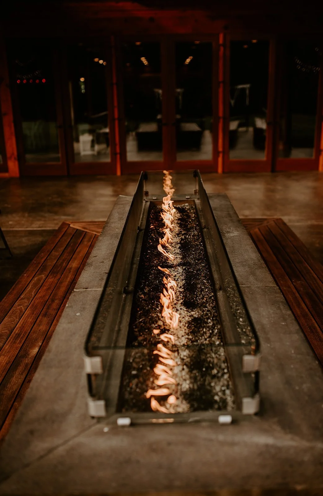 Indoor fire pit with flames on a long, narrow, black bed, surrounded by wooden benches in a cozy, warmly lit room.