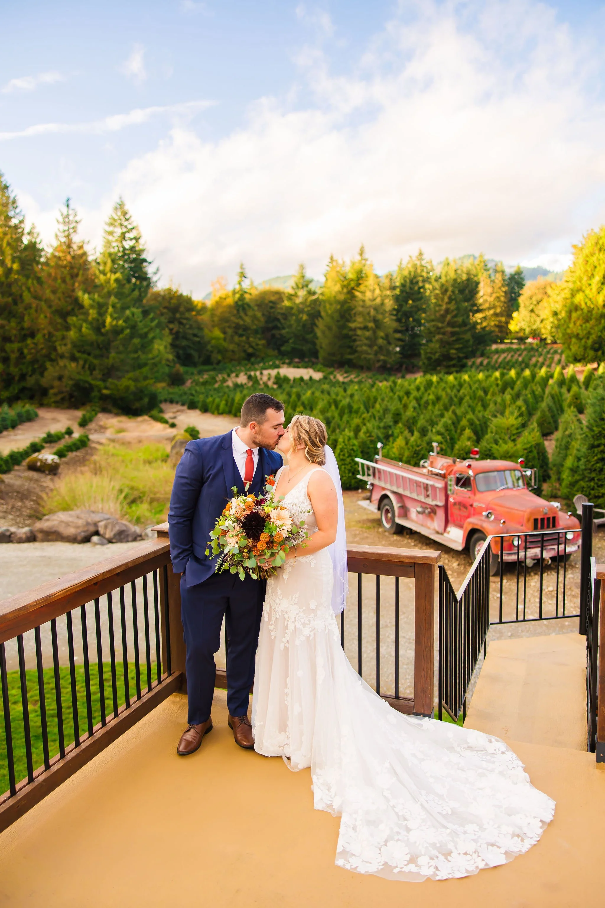 Bride and groom sharing a kiss on a balcony overlooking a vineyard with a red vintage fire truck parked nearby.