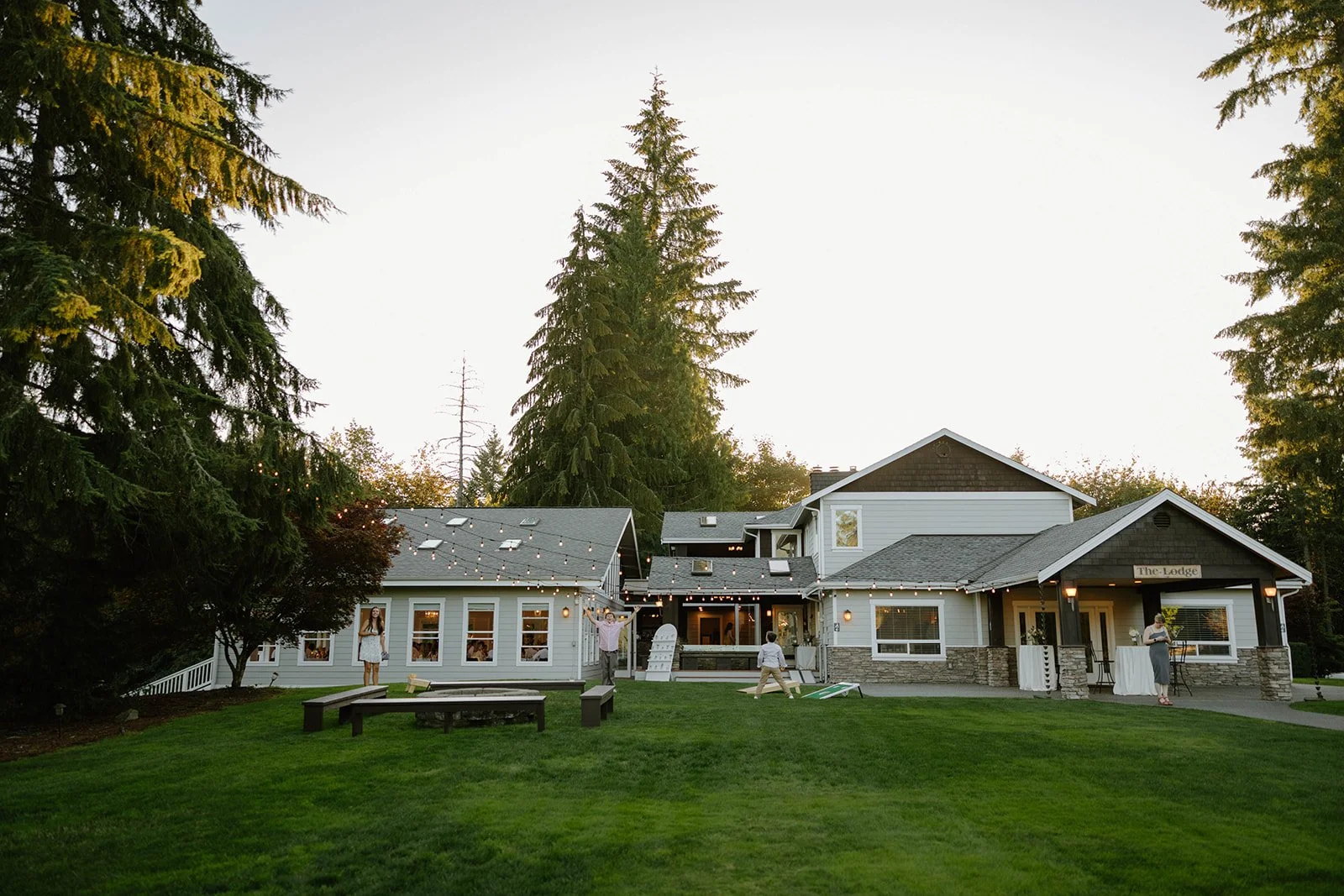 Large house in a wooded area decorated with string lights, with children playing on the lawn and a woman standing near a high table on the porch, during twilight.